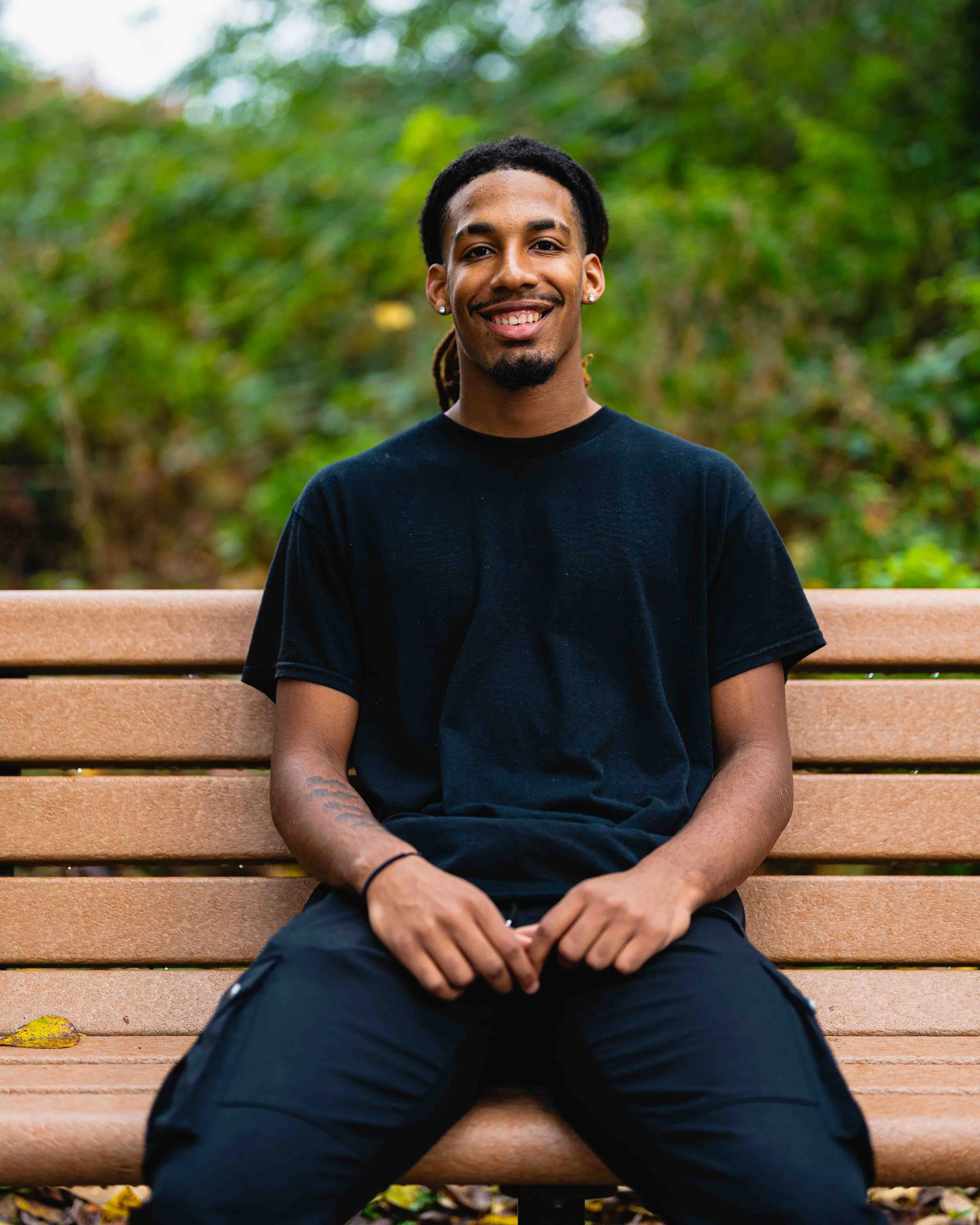 A young man with dark hair, a goatee, earrings, and a black t-shirt sitting on a park bench with green trees in the background, smiling at the camera.