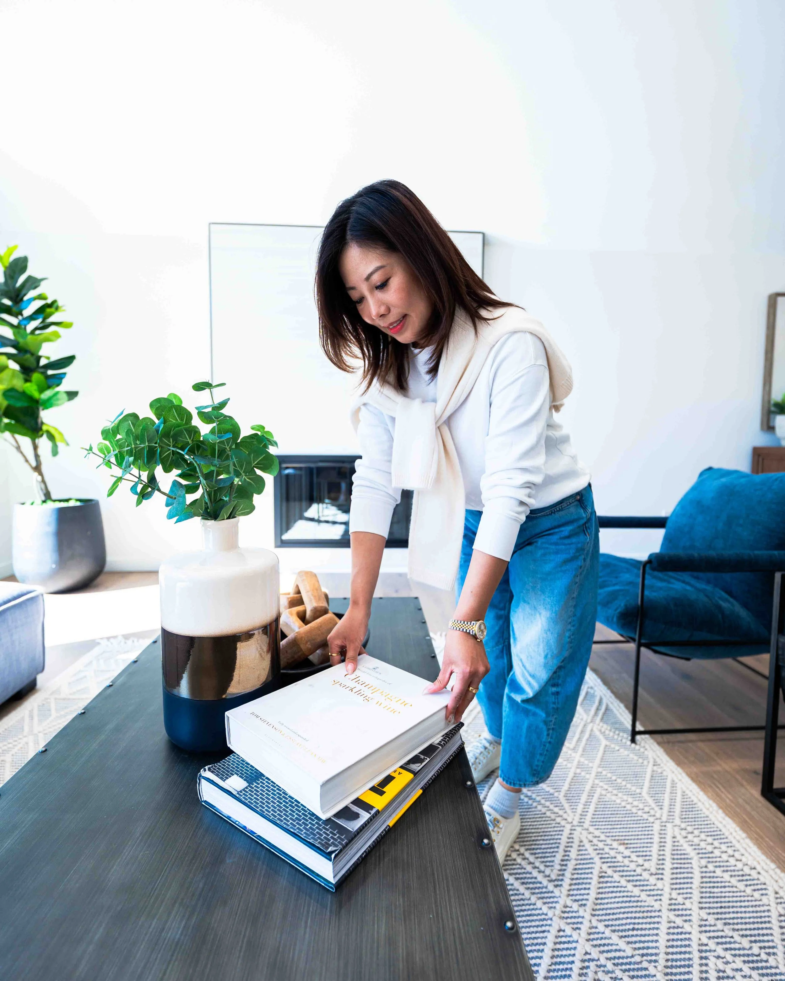 Woman leaning over a table arranging books and a box, with potted plants and a sofa in the background.