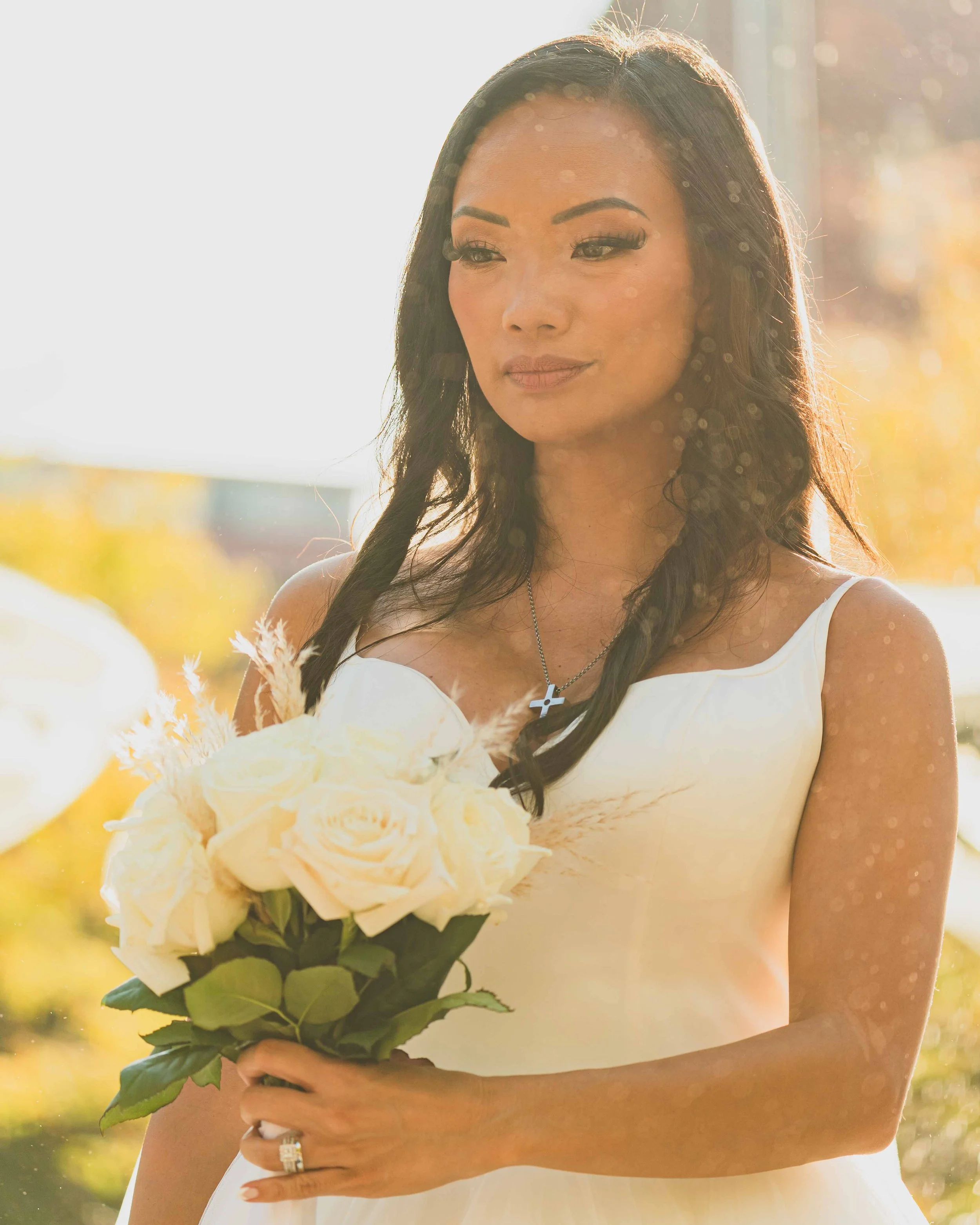 A woman with long dark hair, wearing makeup, a white dress, and a cross necklace, is holding a bouquet of cream-colored roses with green leaves. She is outdoors in sunlight with a blurred background of yellow and green hues, suggesting a sunny day.