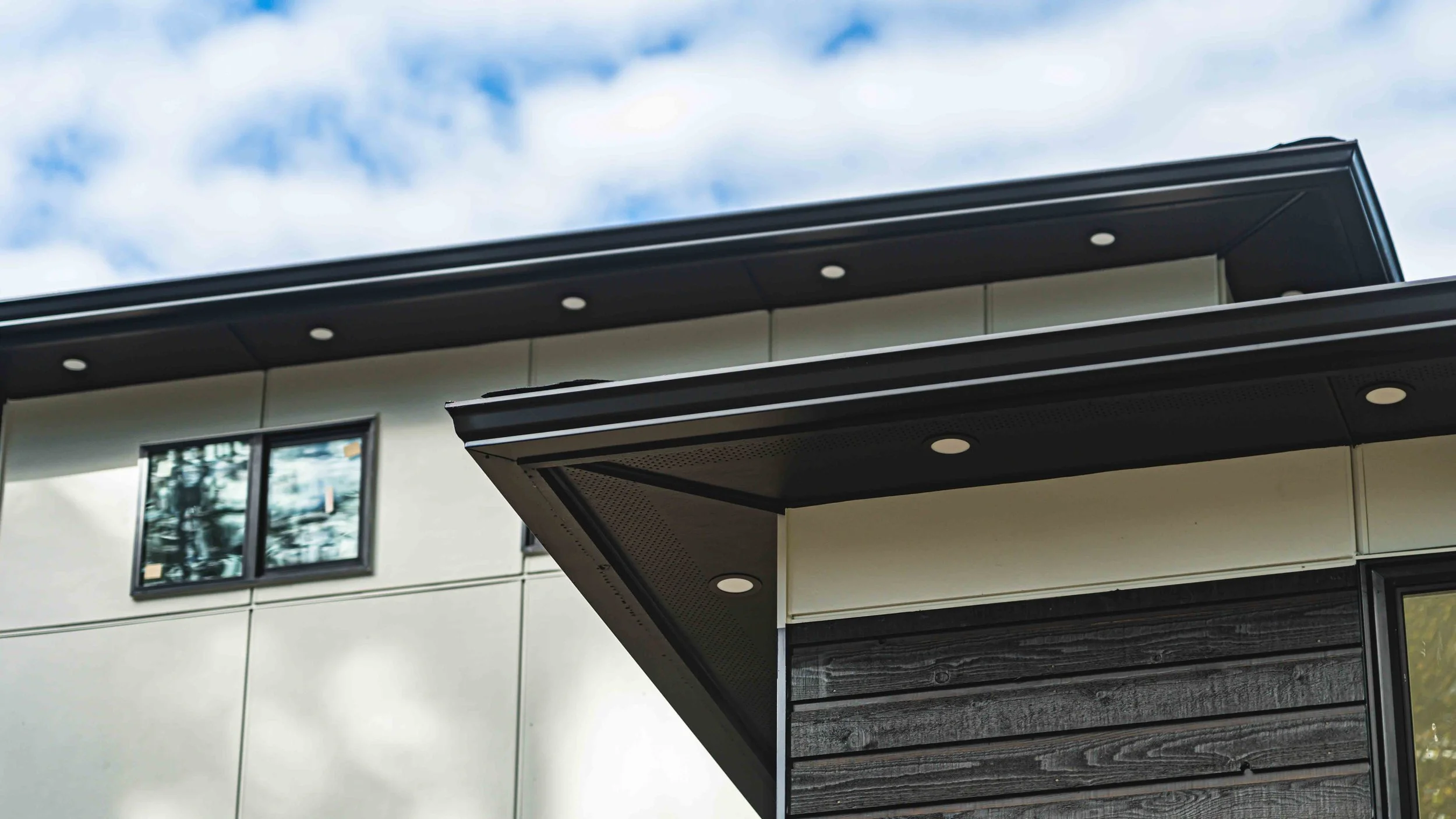 Close-up of modern building's exterior with black roof eaves, recessed lighting, large window, and light-colored siding, under a cloudy sky.