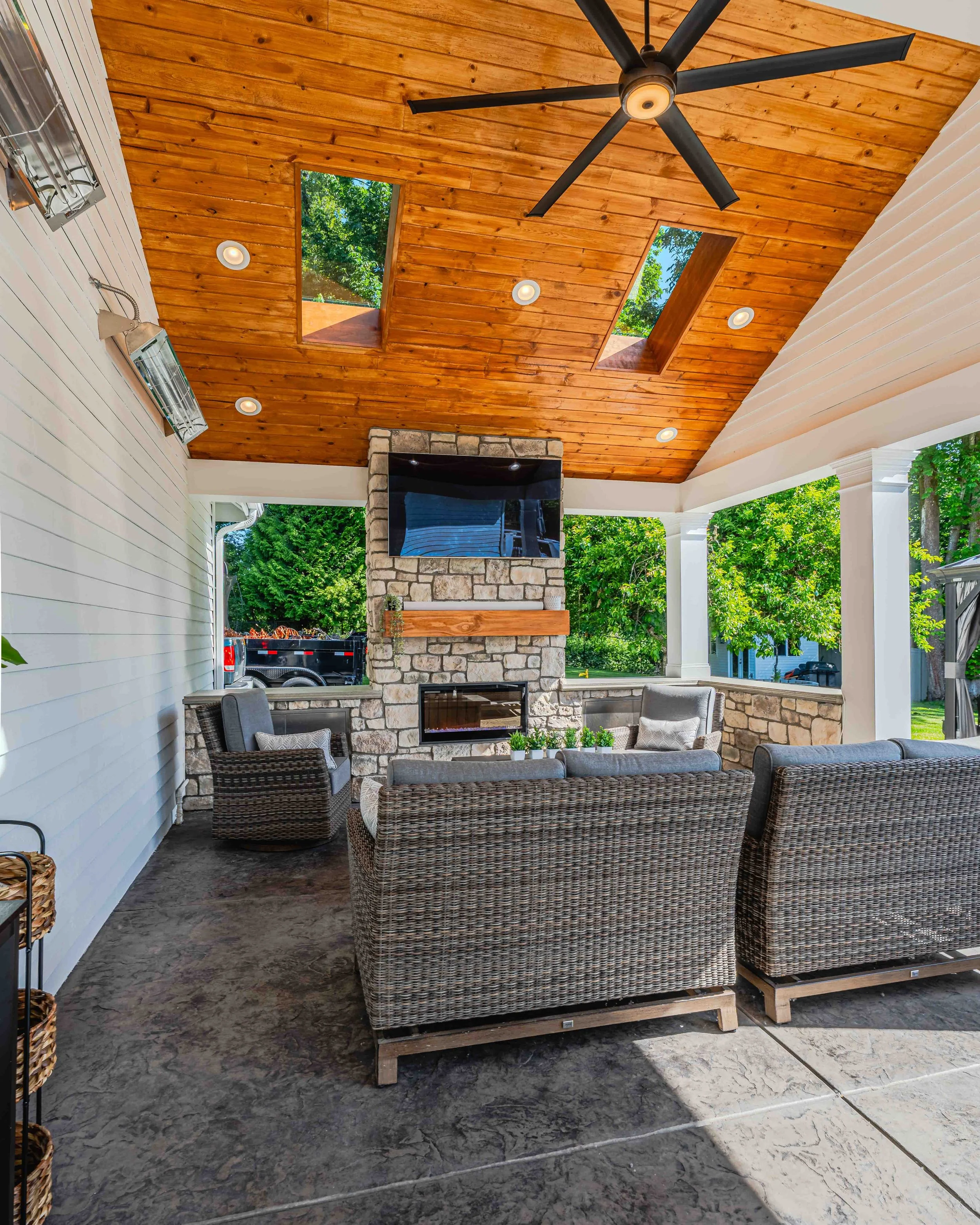 Covered outdoor patio with a wood-paneled ceiling featuring skylights, ceiling fan, stone fireplace with a TV, wicker chairs, and a marble floor.