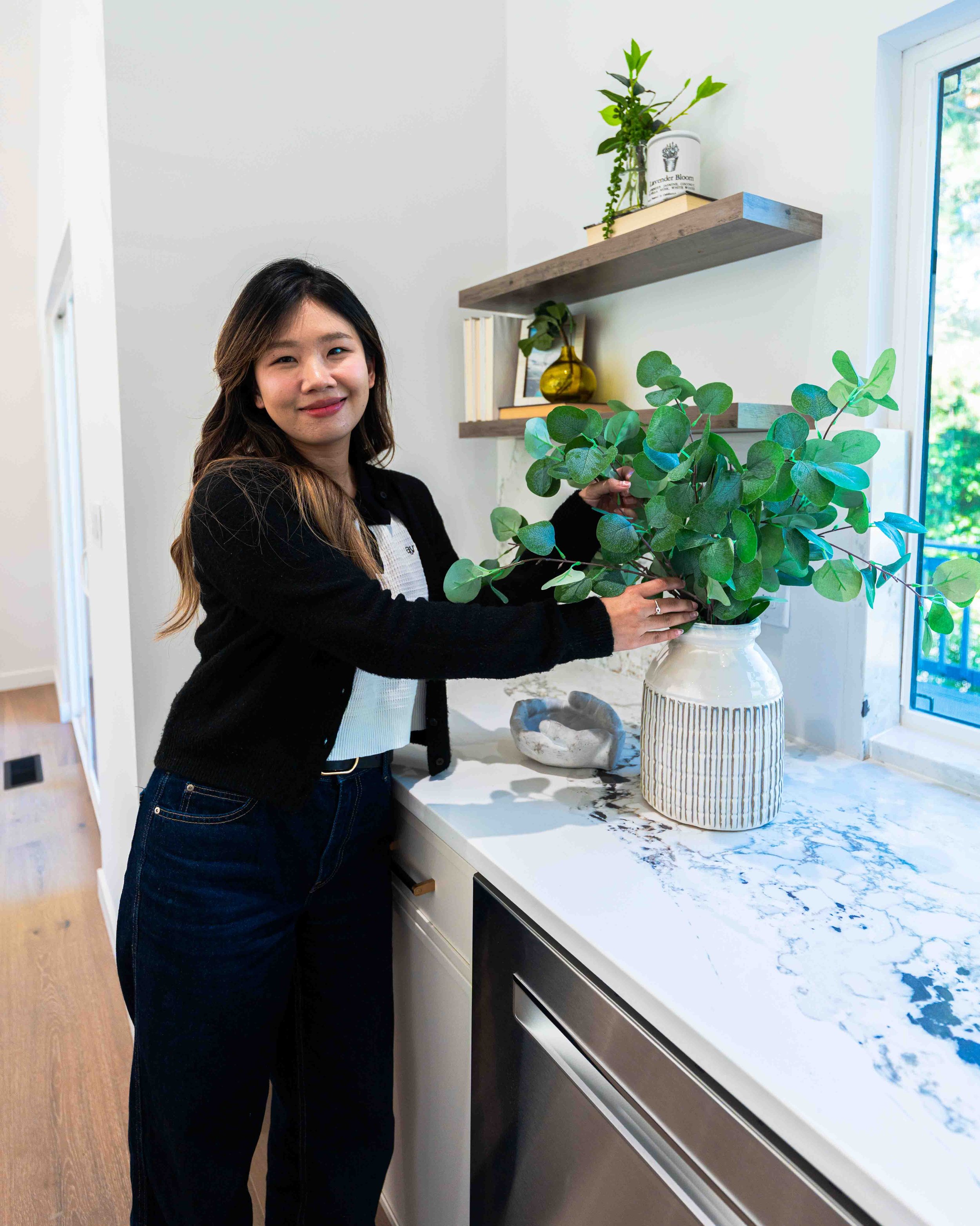 A woman with long dark hair, wearing a black sweater and jeans, is placing a potted green plant on a white marble kitchen countertop near a window. Two wooden shelves with decorative items and books are on the white wall behind her.