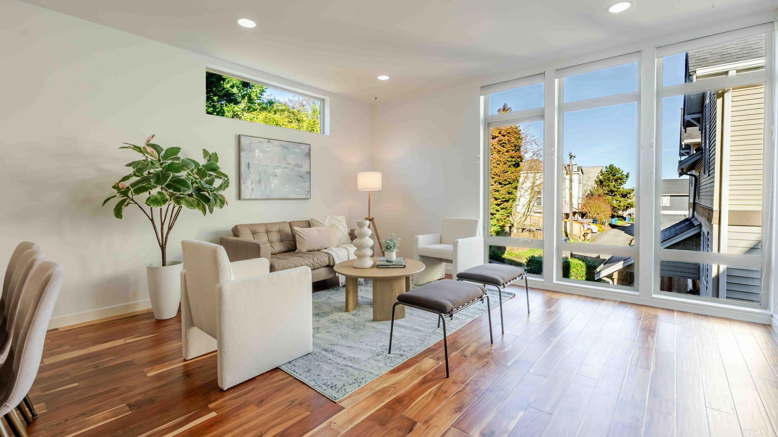 Bright living room with large windows showing outdoor view, wood floors, beige couches, white armchair, decorative plant, wall art, and a side table with a lamp.