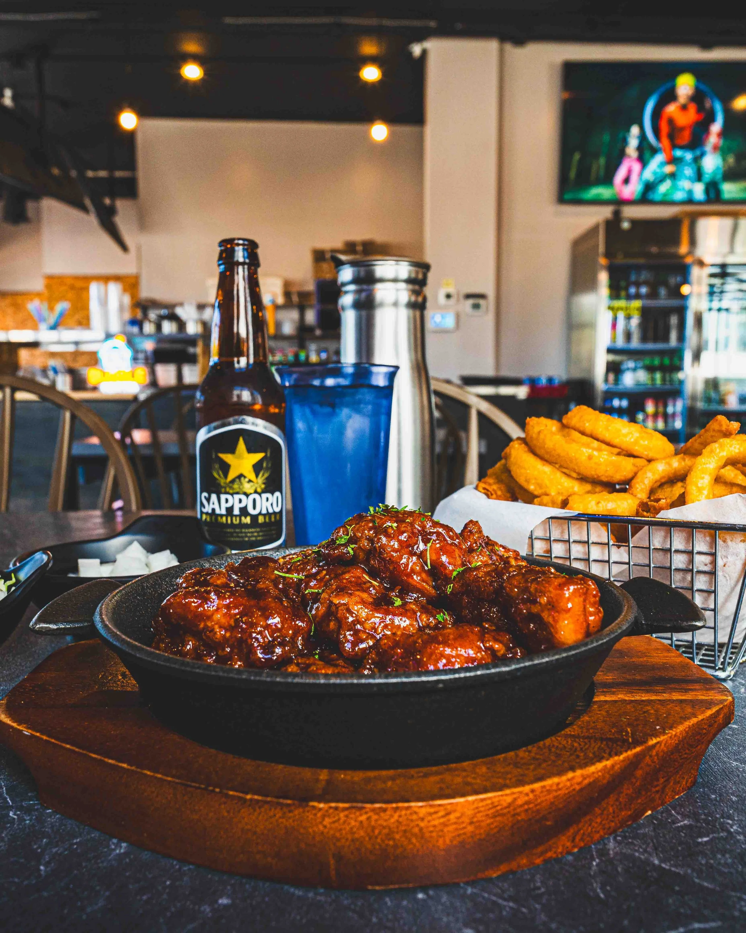 Close-up of a spicy glazed chicken dish served in a black skillet on a wooden tray at a restaurant. In the background, there is a plate of fried onion rings, a bottle of Sapporo beer, a blue cup, and a countertop. The restaurant interior includes a t
