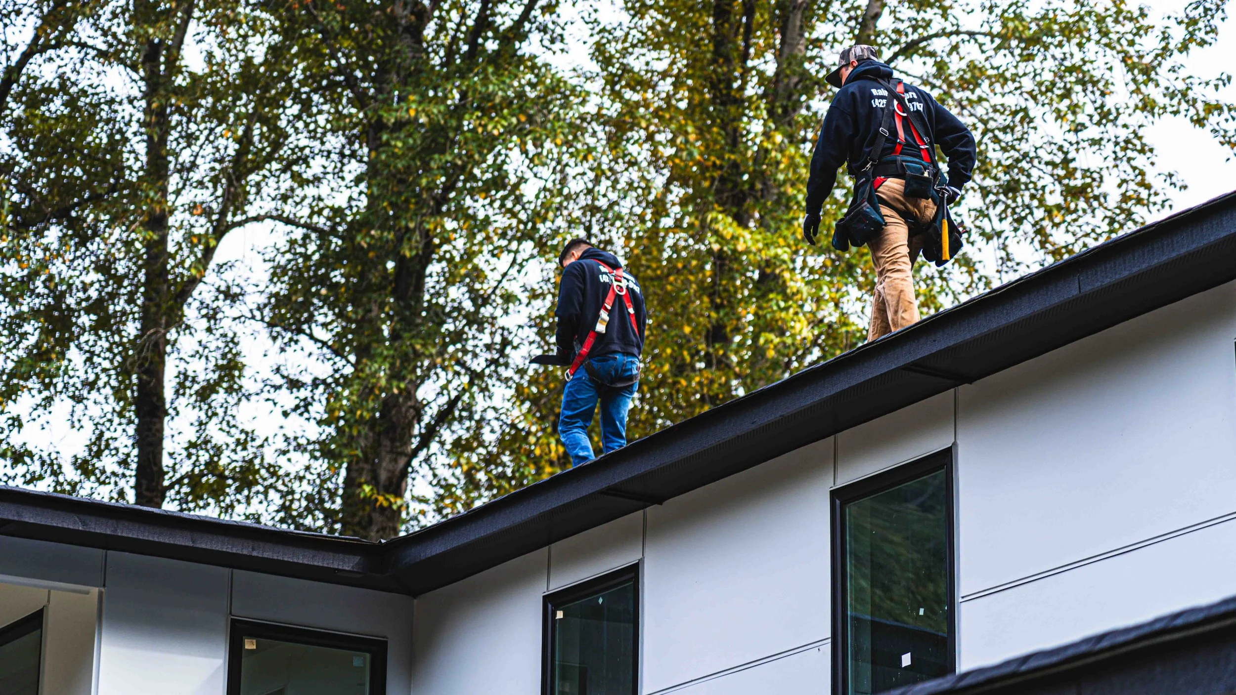 Two workers wearing safety harnesses on the roof of a modern residential building, with a background of tall trees with green and yellow leaves.