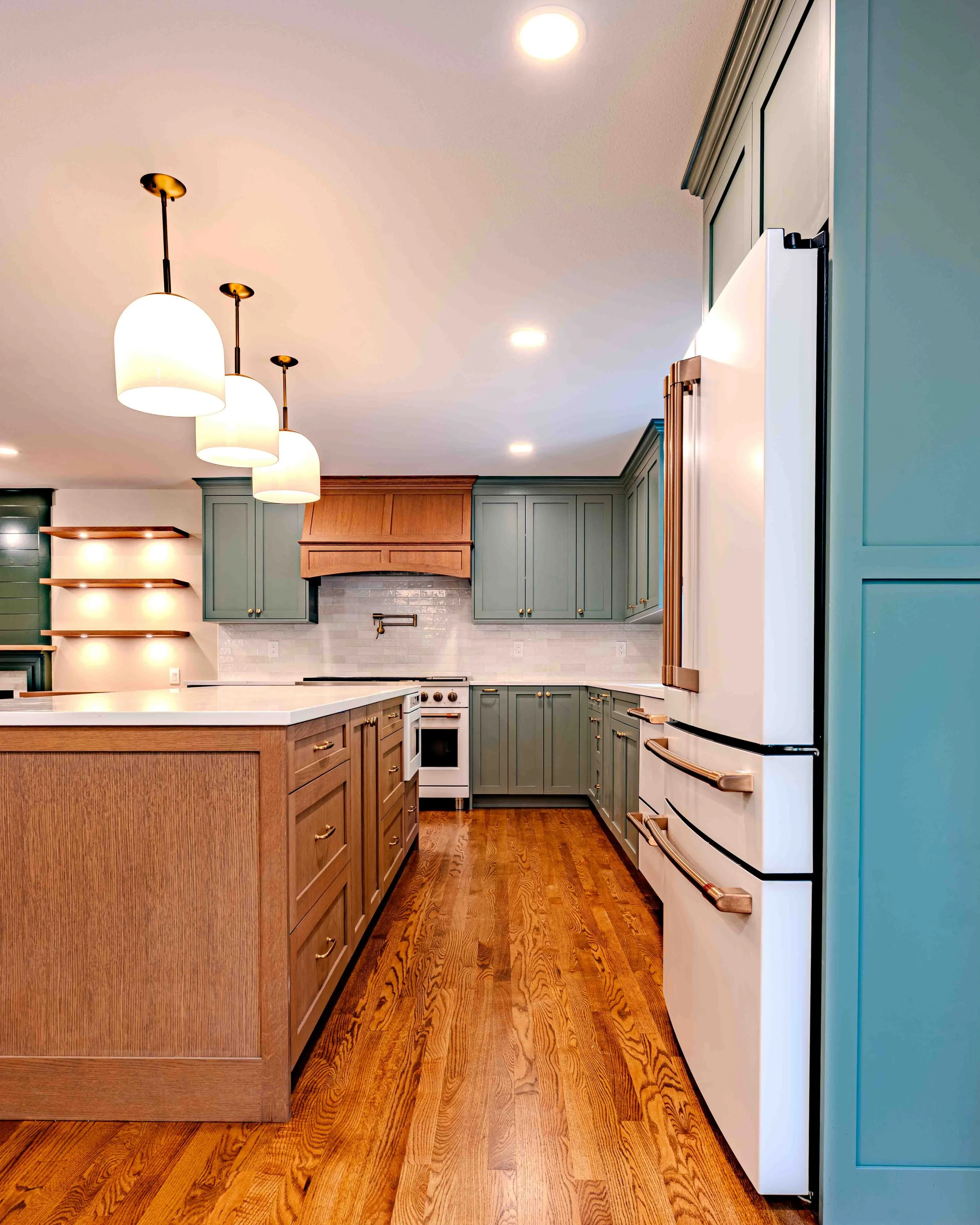 Modern kitchen with green cabinetry, a white refrigerator with gold handles, a white fireplace, and a wooden kitchen island with drawers. Pendant lights hang from the ceiling.