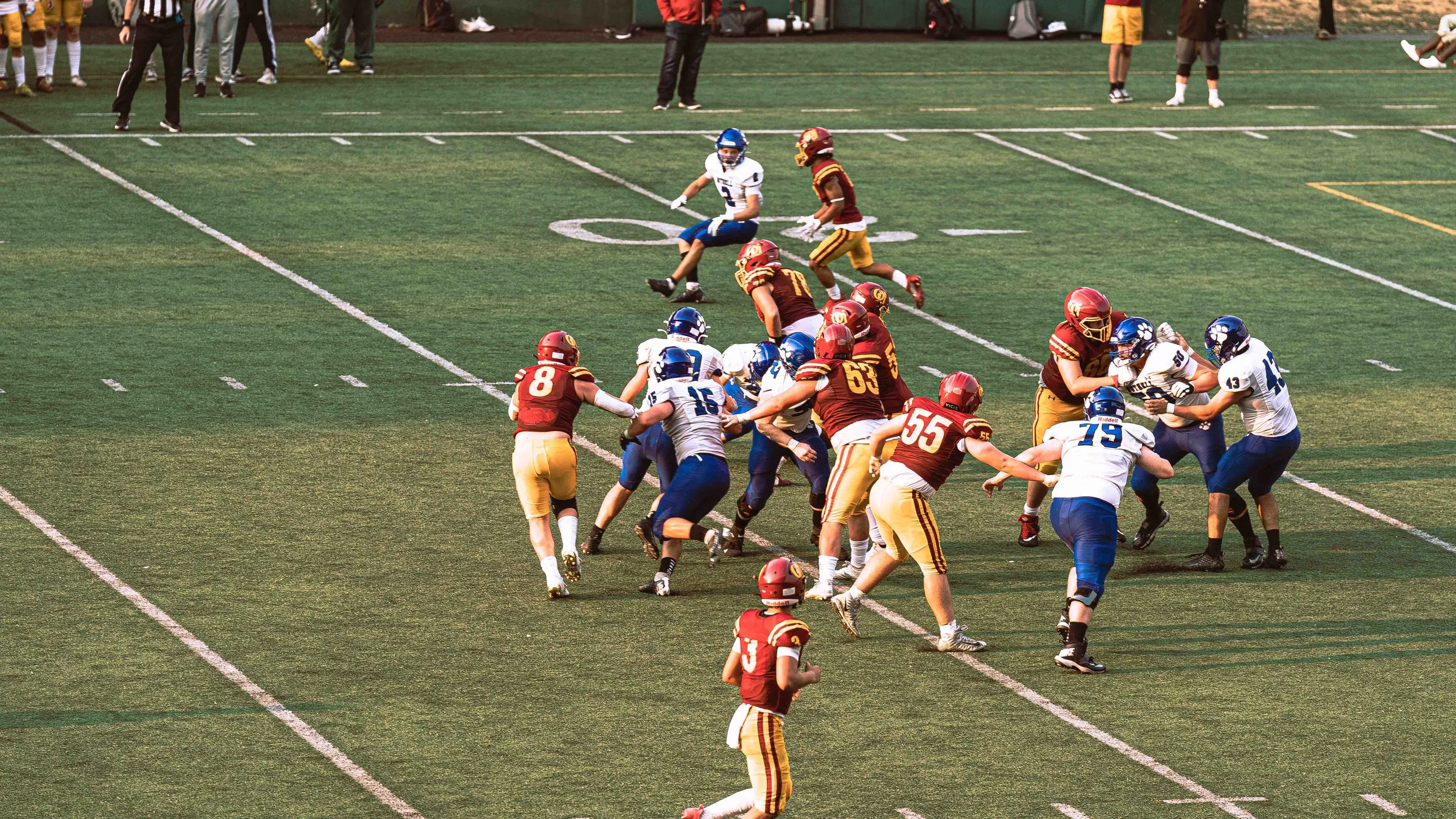 A youth football game on a green field, with players in red and yellow uniforms and players in white and blue uniforms, actively engaged in a play, with some players tackling and others running.