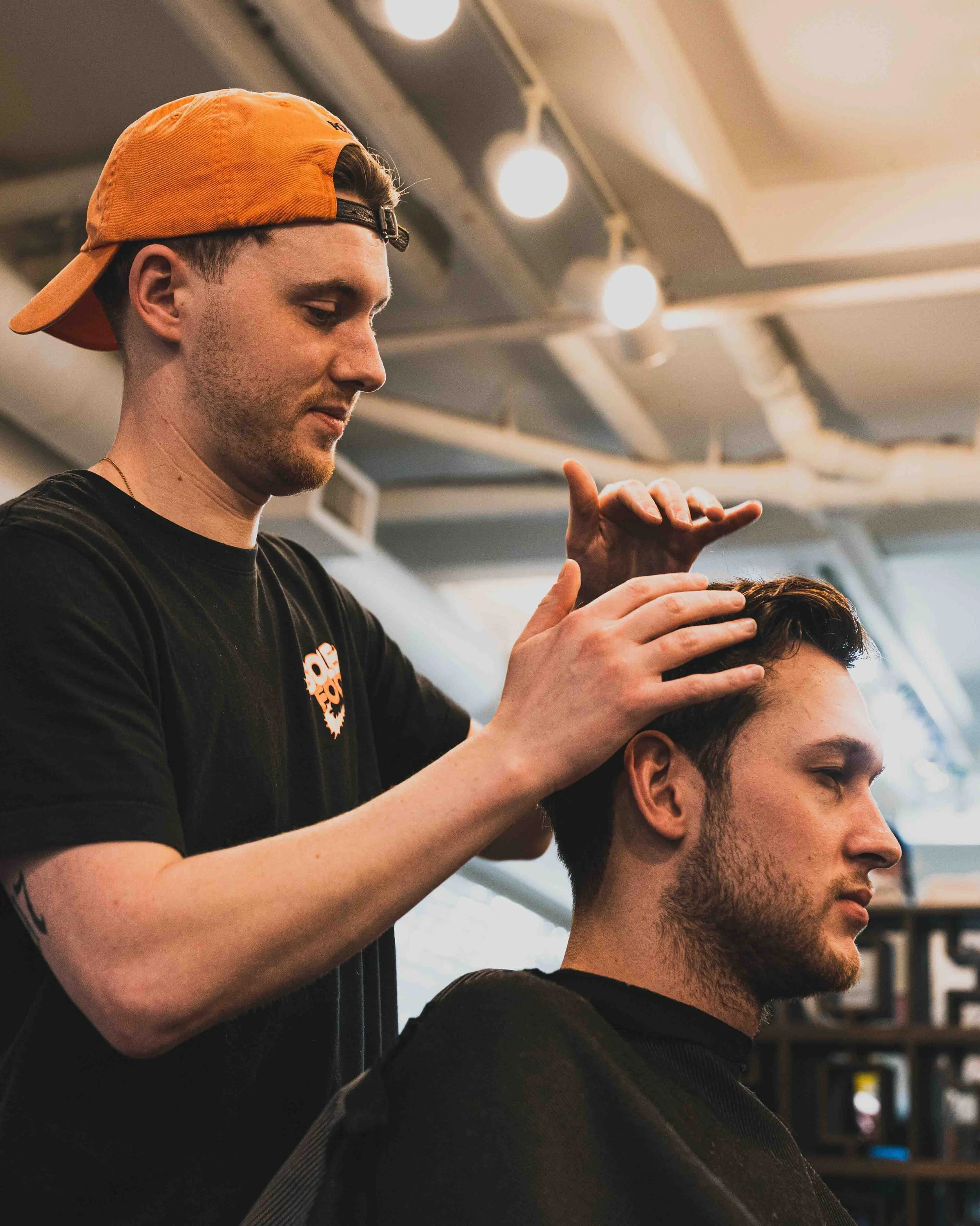 A hairstylist wearing an orange cap and black shirt is giving a haircut to a male client with dark hair and a beard in a salon with ceiling lights.