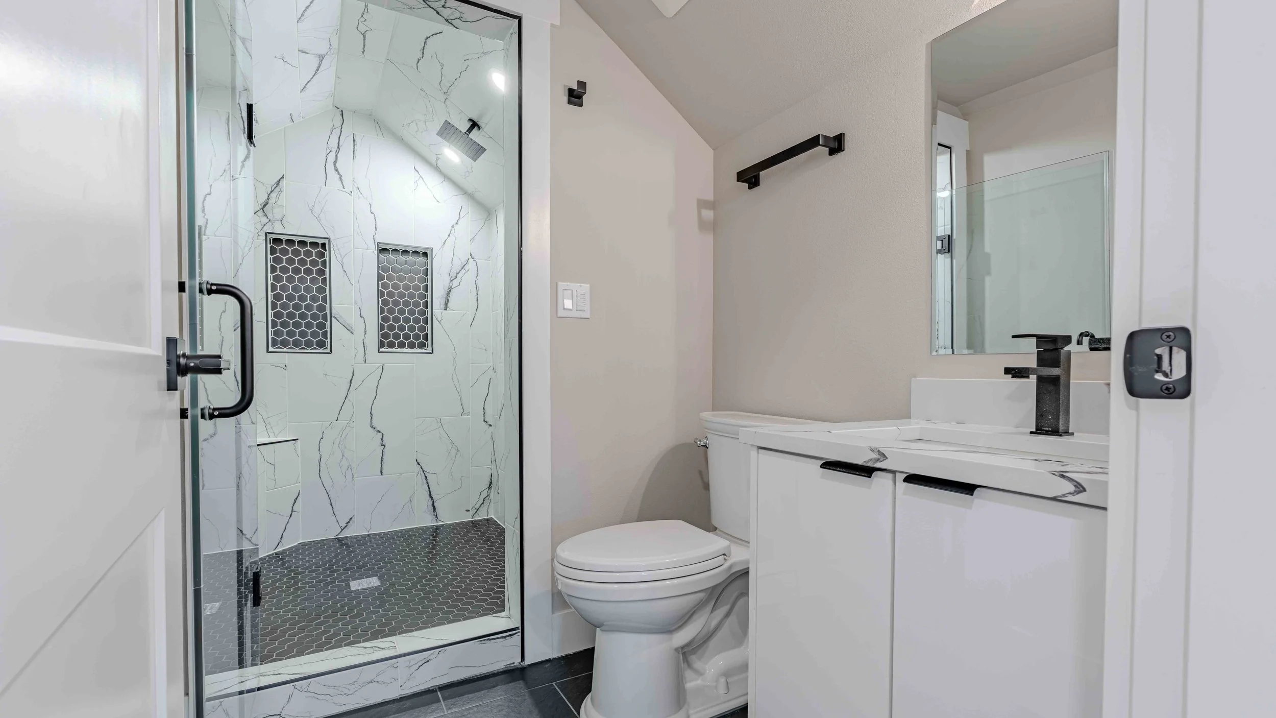 Modern bathroom with a marble vanity, black fixtures, a large mirror, a toilet, and a walk-in shower with marble walls and black hexagonal floor tiles.