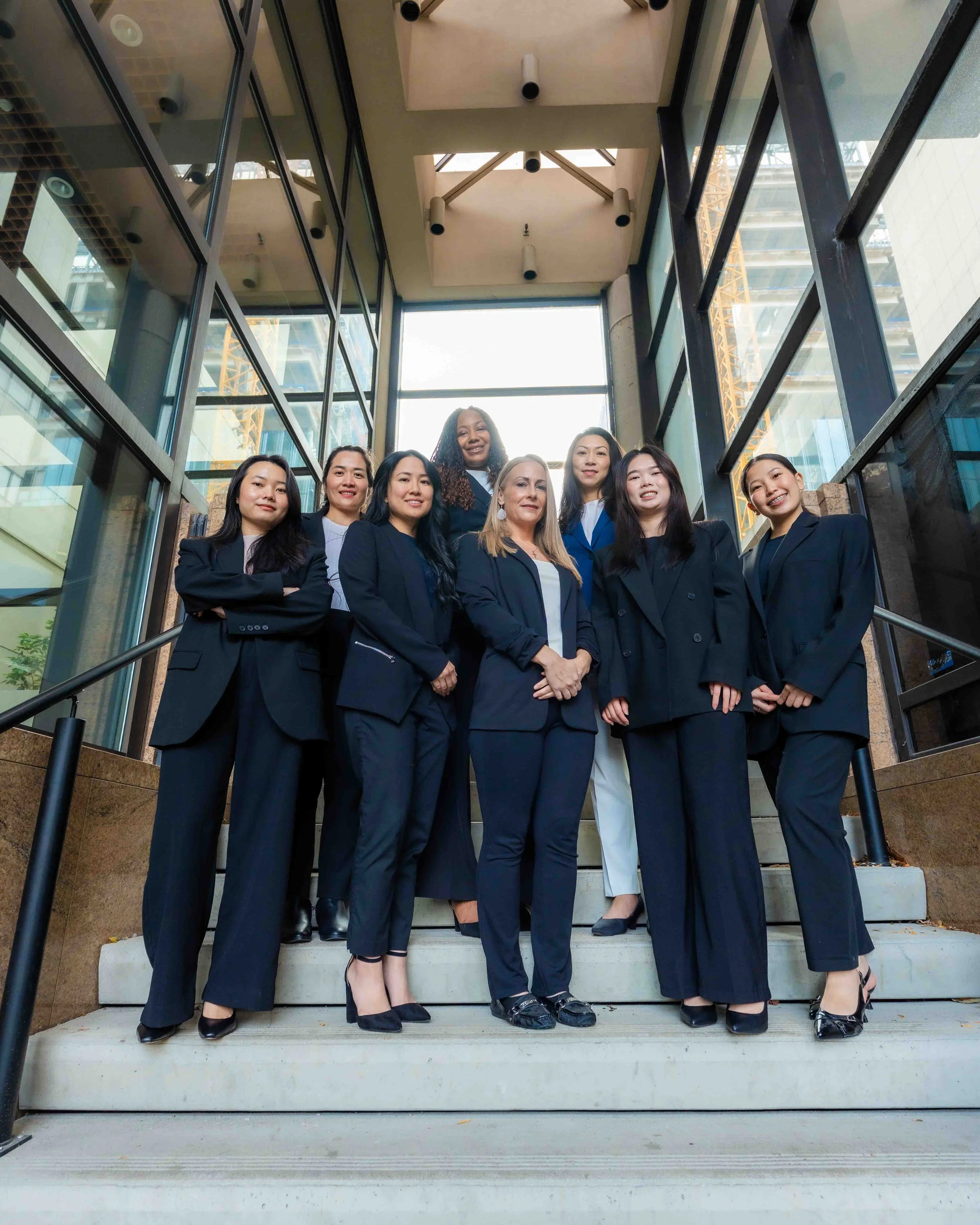 A group of seven women dressed in formal business attire standing on outdoor steps inside a modern building with glass walls and metal framing, smiling at the camera.