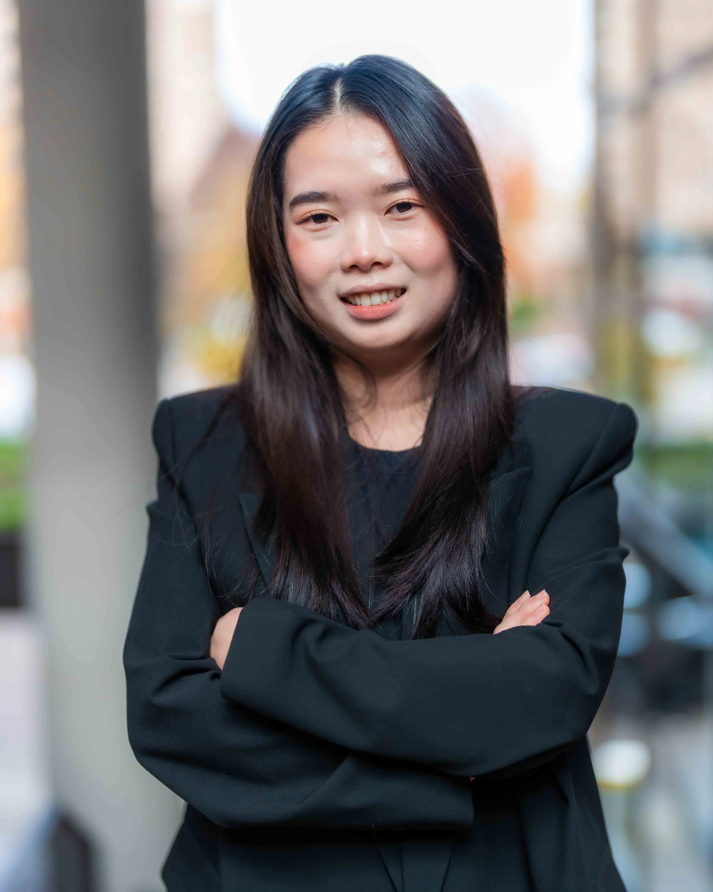 Portrait of a smiling Asian woman with long dark hair, wearing a black blazer, standing outdoors with a blurred cityscape background.