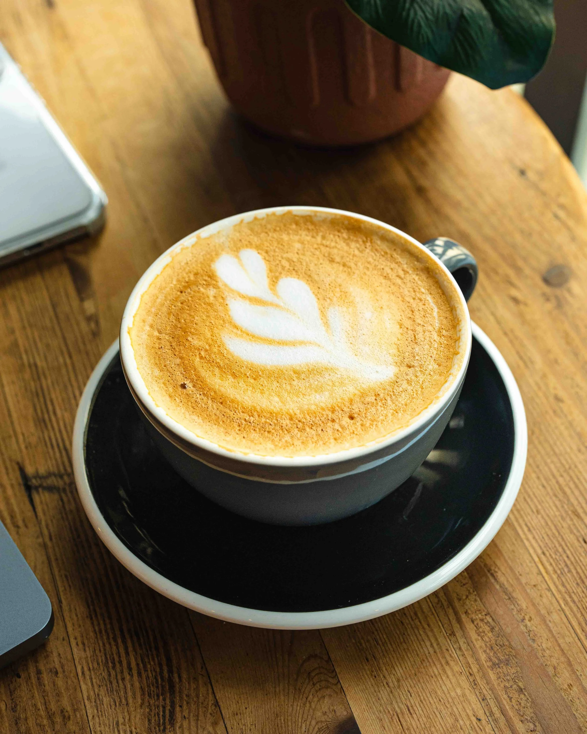 A cup of coffee with latte art on top, placed on a black saucer on a wooden table, with a potted plant nearby.