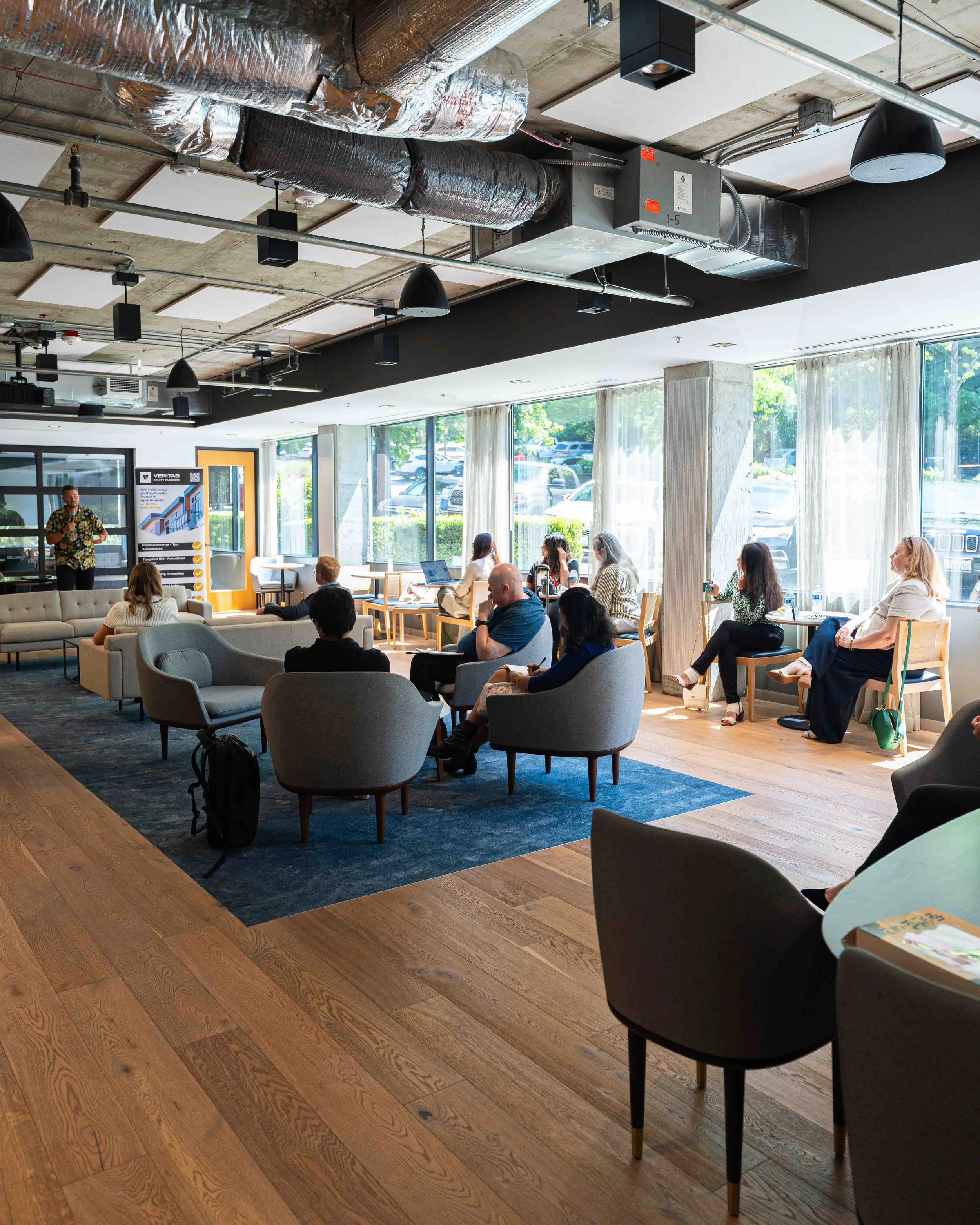 A modern indoor meeting or conference room with large windows, wood flooring, and a mix of lounge and dining seating. There are people sitting and listening to a person presenting at the front. The ceiling features exposed ductwork, vents, and black 