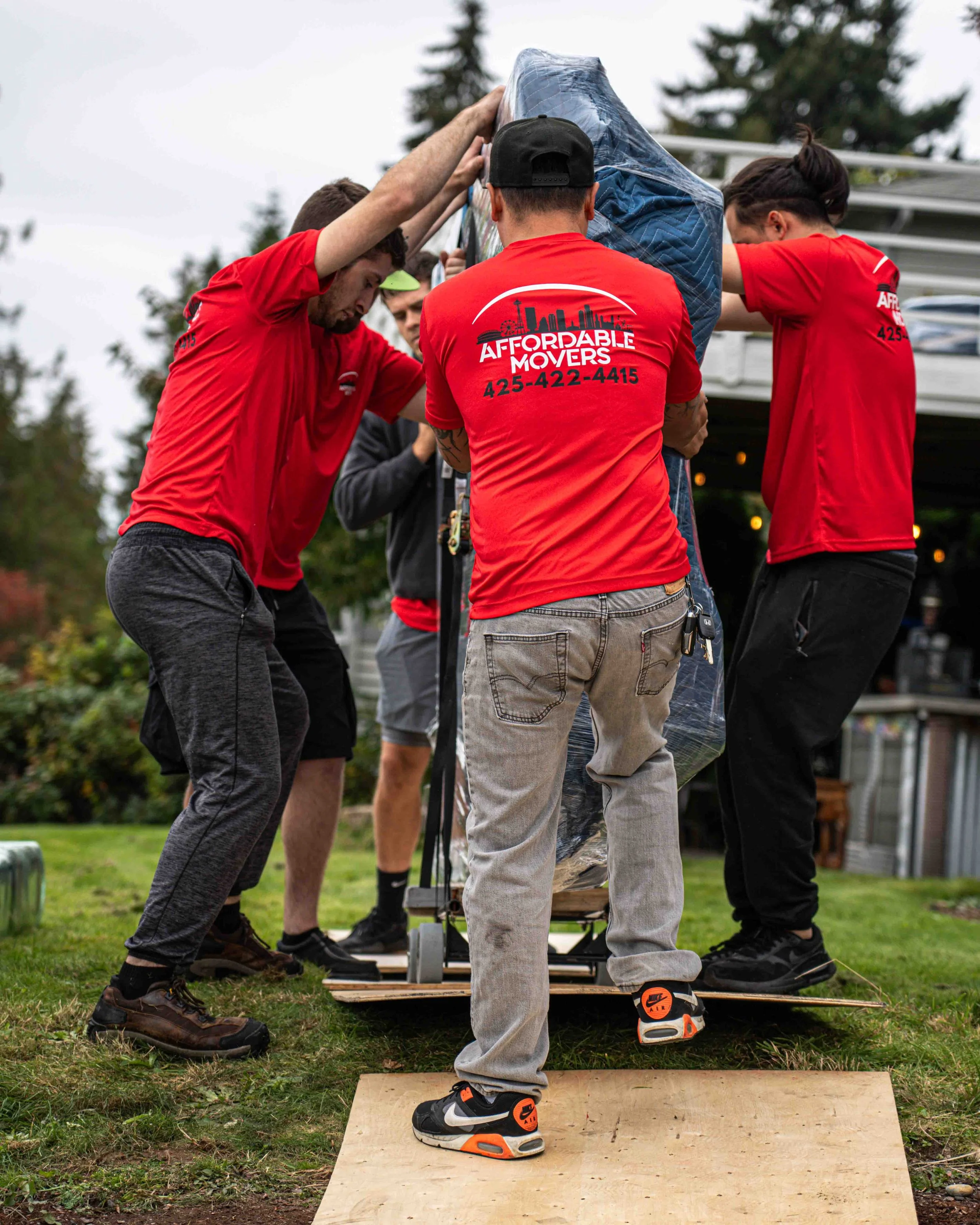 A group of people working together outdoors on a construction or moving project, wearing red shirts with company branding, on a makeshift platform with a wooden board on grass, with trees and a building in the background.