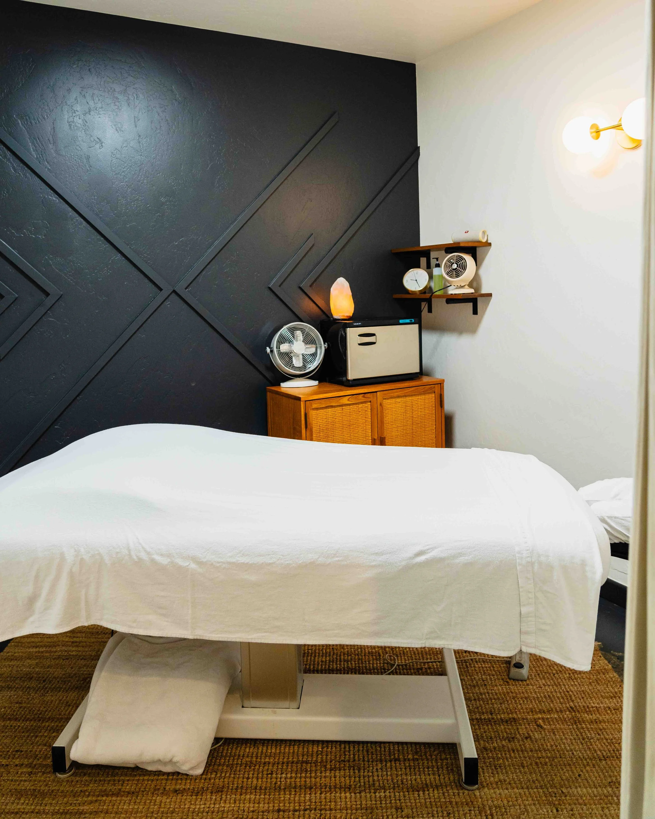 Hospital bed with white sheets and pillow, partially covered by white blanket, in a room with dark accent wall, wooden cabinet, standing fan, salt lamp, small shelf with clock, lamp, and paper roll, beige carpeted floor.