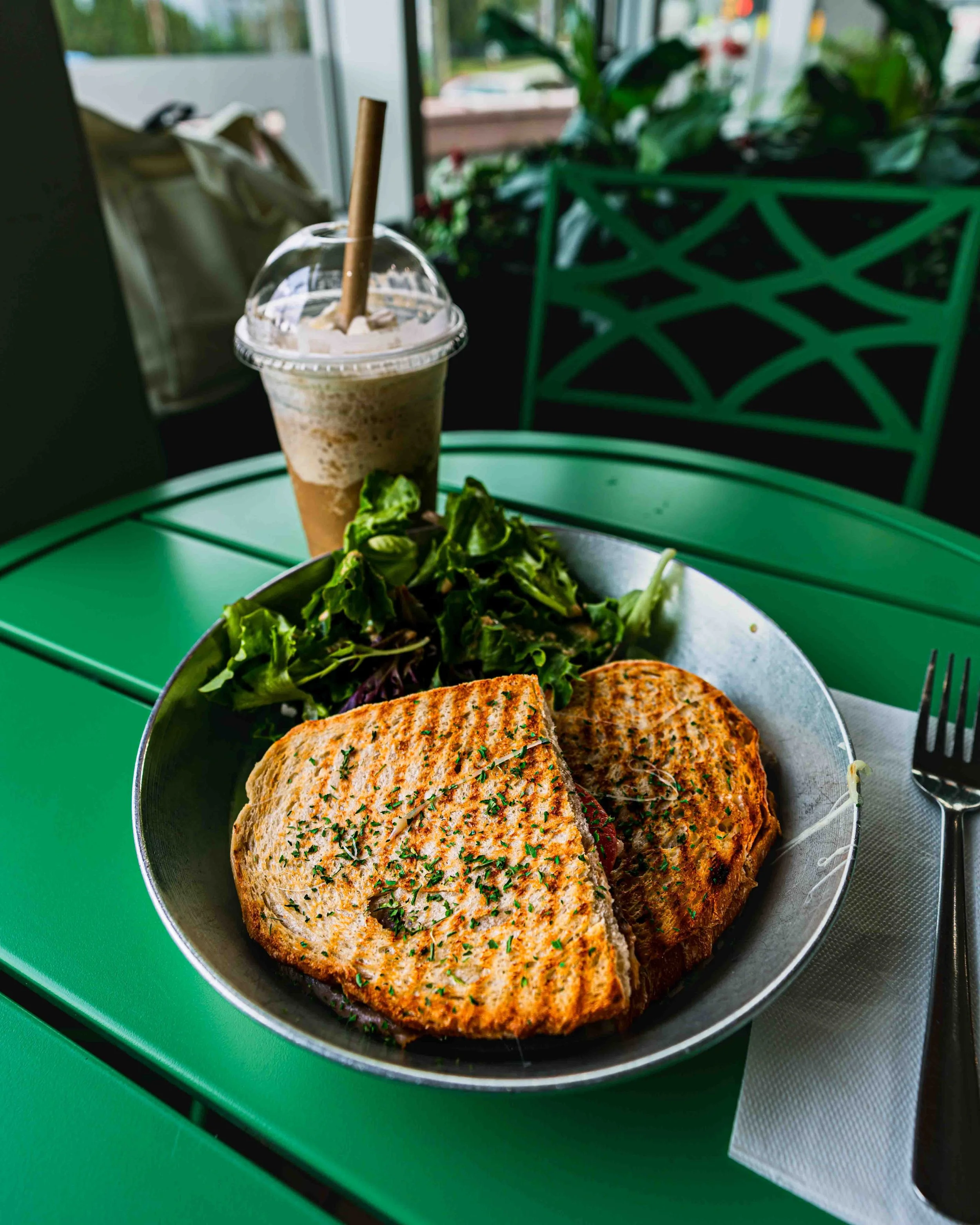 A grilled chicken panini sandwich with herbs on toasted bread, served with a side salad, on a metal plate; a caramel frappuccino with whipped cream in a plastic cup with a lid and straw, on a green table near a green chair with plants and window in t