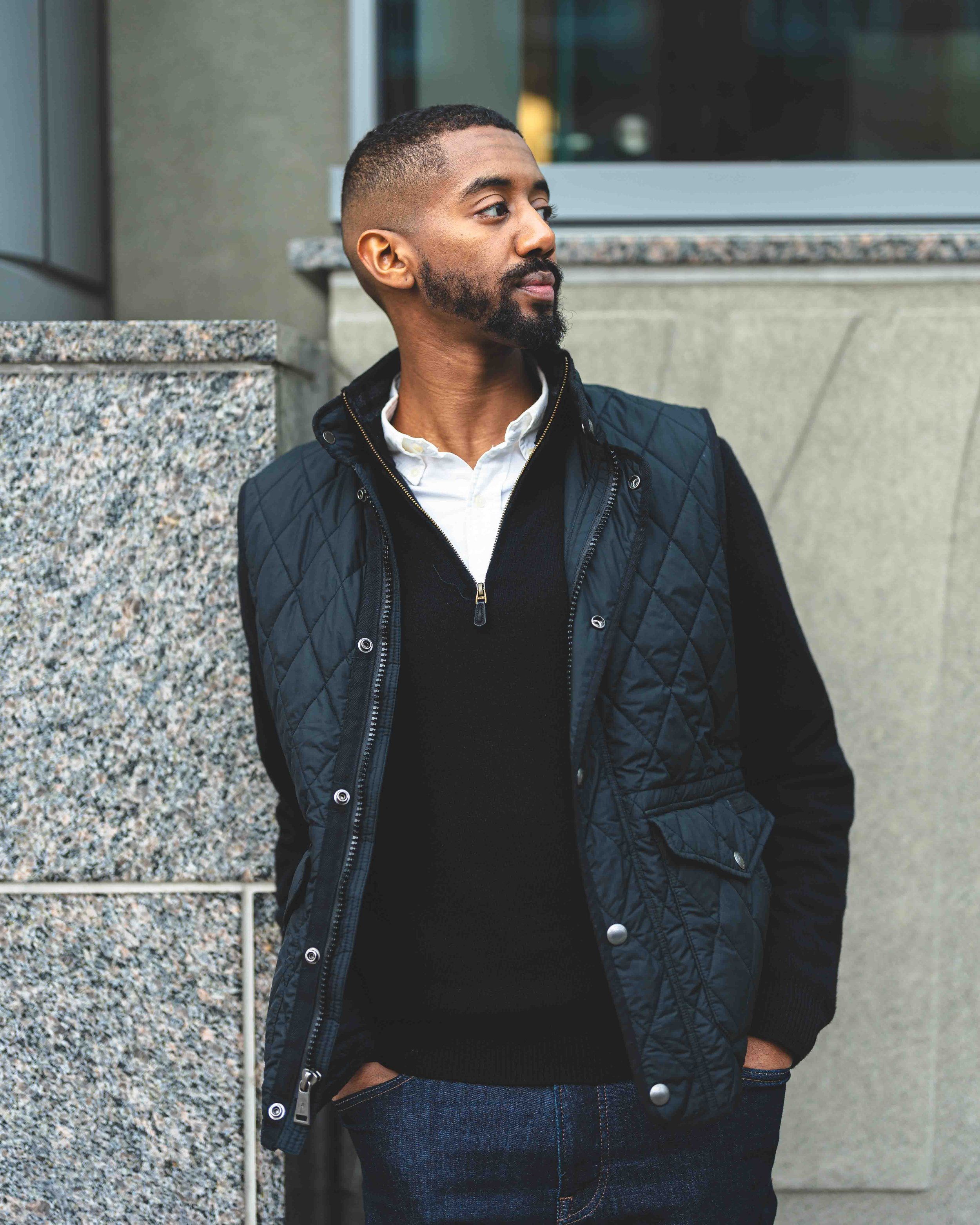 A man standing outdoors with his hands in his pockets, wearing a black quilted jacket over a white shirt and a black sweater, looking to his right, with a building and a large window in the background.