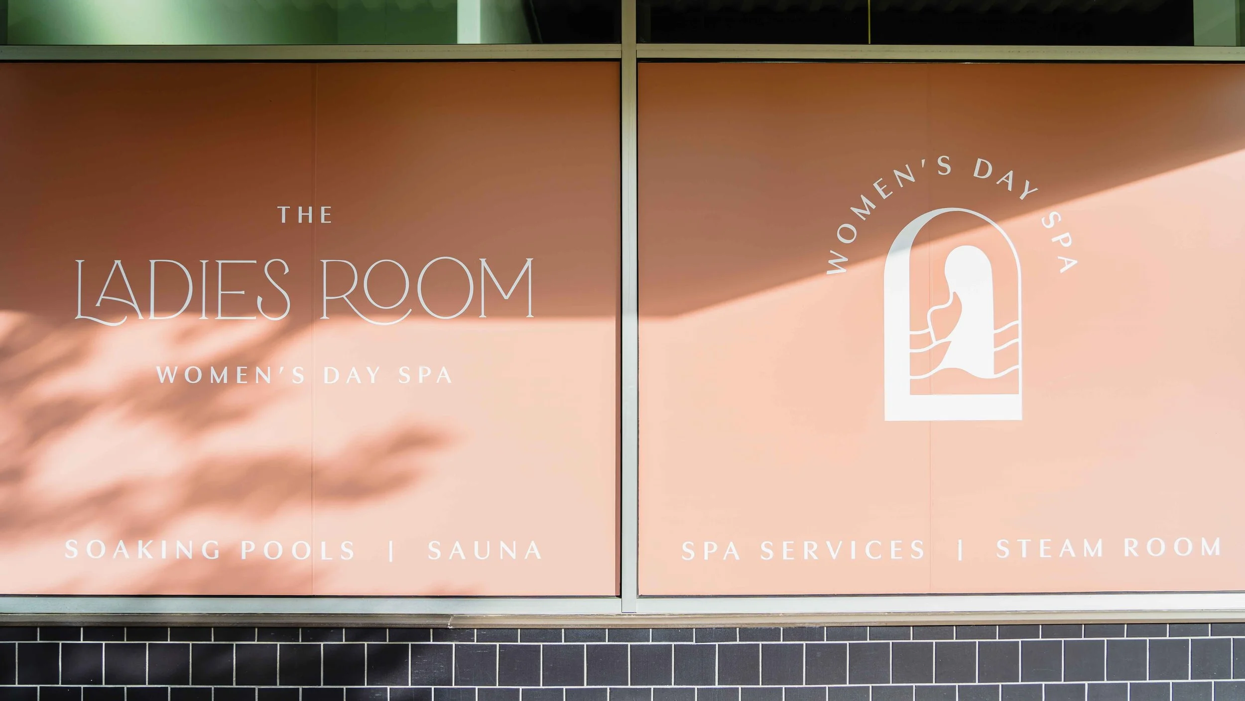 Sign for a women's day spa called The Ladies Room, featuring a stylized outline of a woman's face and hair, with services listed below, including soaking pools, sauna, spa services, and steam room.