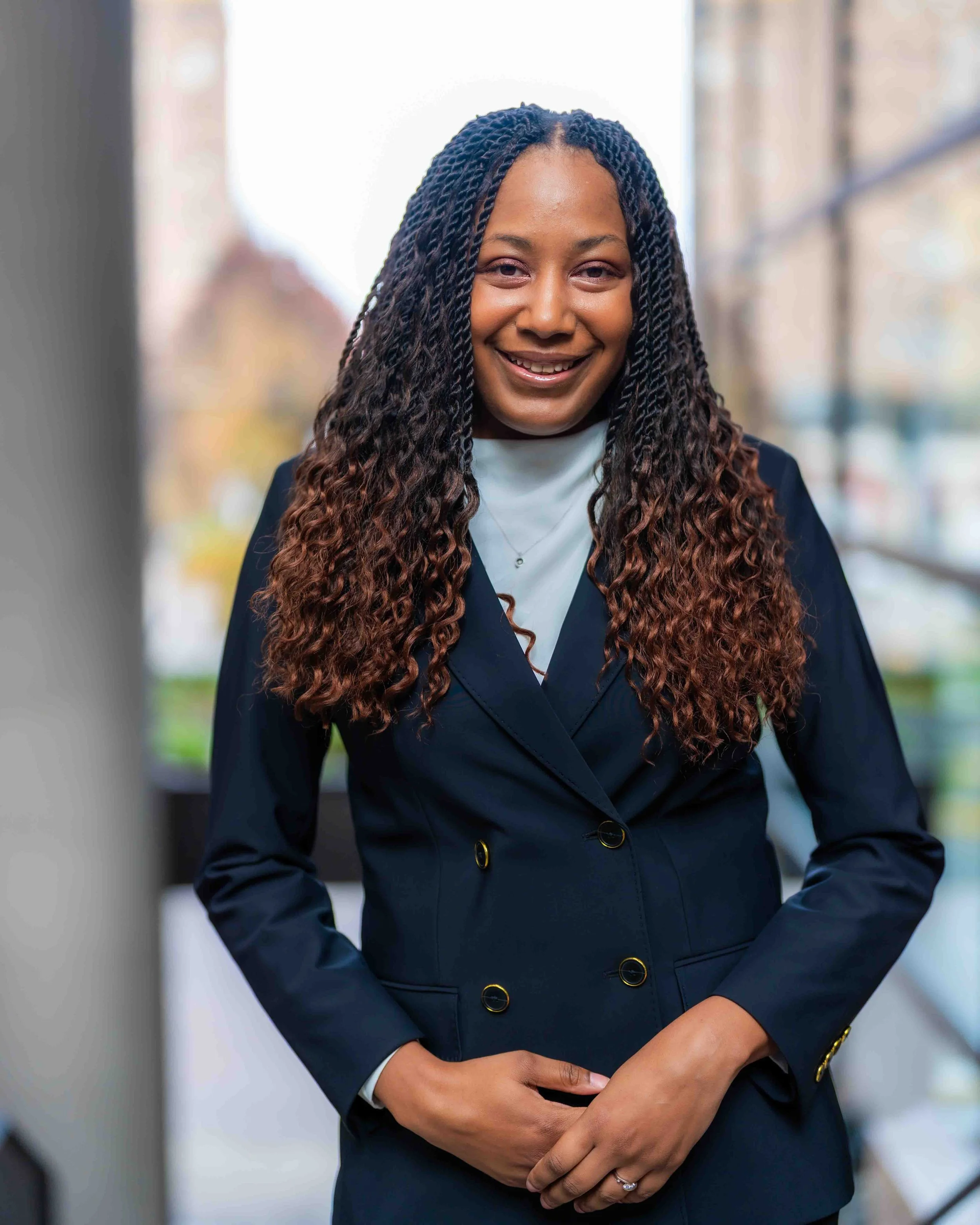 A woman with long curly hair, smiling, wearing a navy blazer and white blouse, standing outdoors with blurred background of buildings and trees.