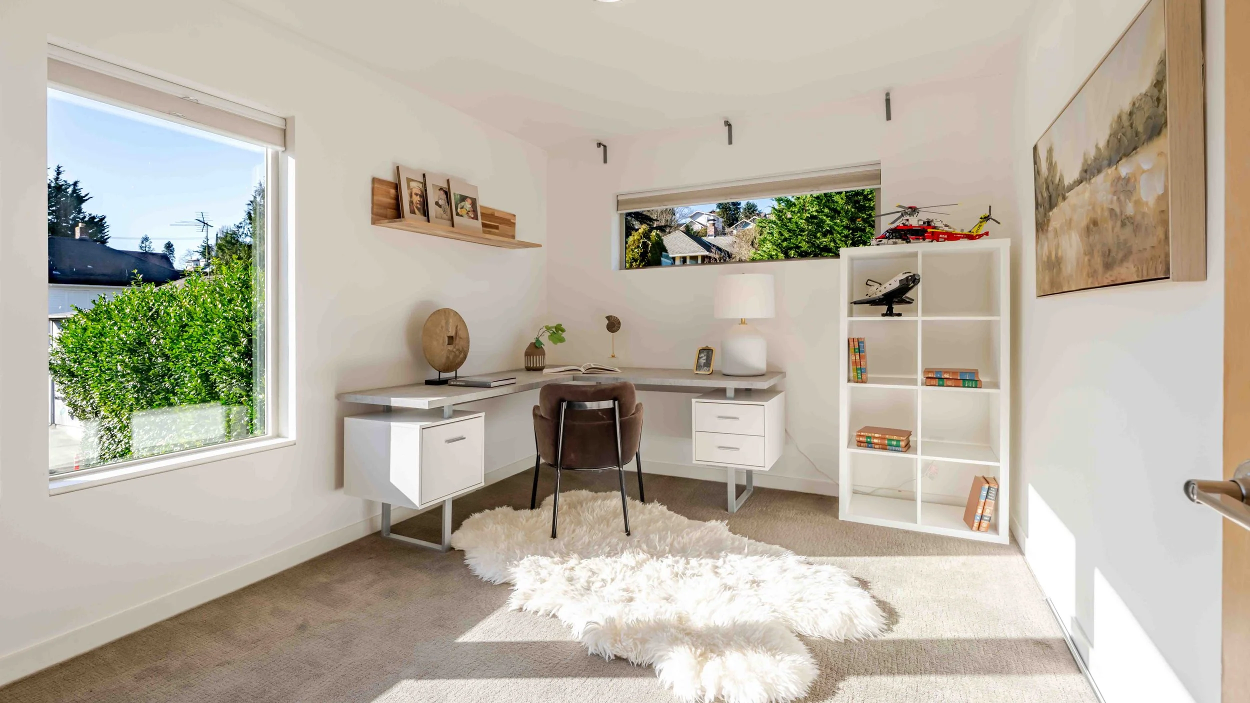 A bright home office with a large window, white walls, a desk with a chair, a white bookshelf with books and toys, and outdoor greenery visible through the window.