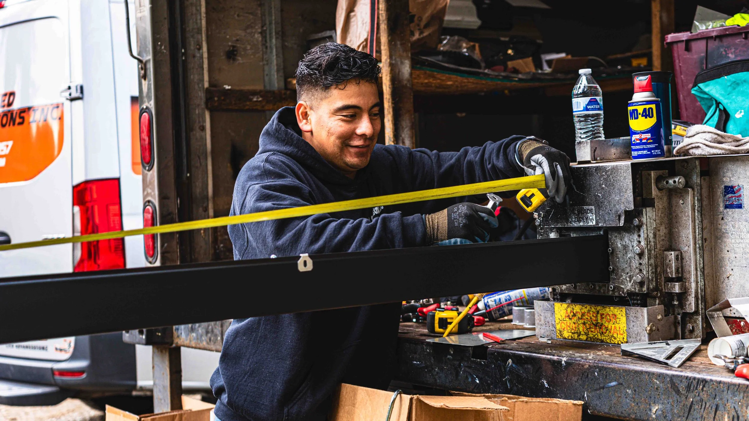 A young man with short, curly dark hair, wearing a dark hoodie and work gloves, is working on a device or machine in a workshop. He is smiling while using a tool, with work supplies, bottles, a water container, and tools around him. There is a yellow