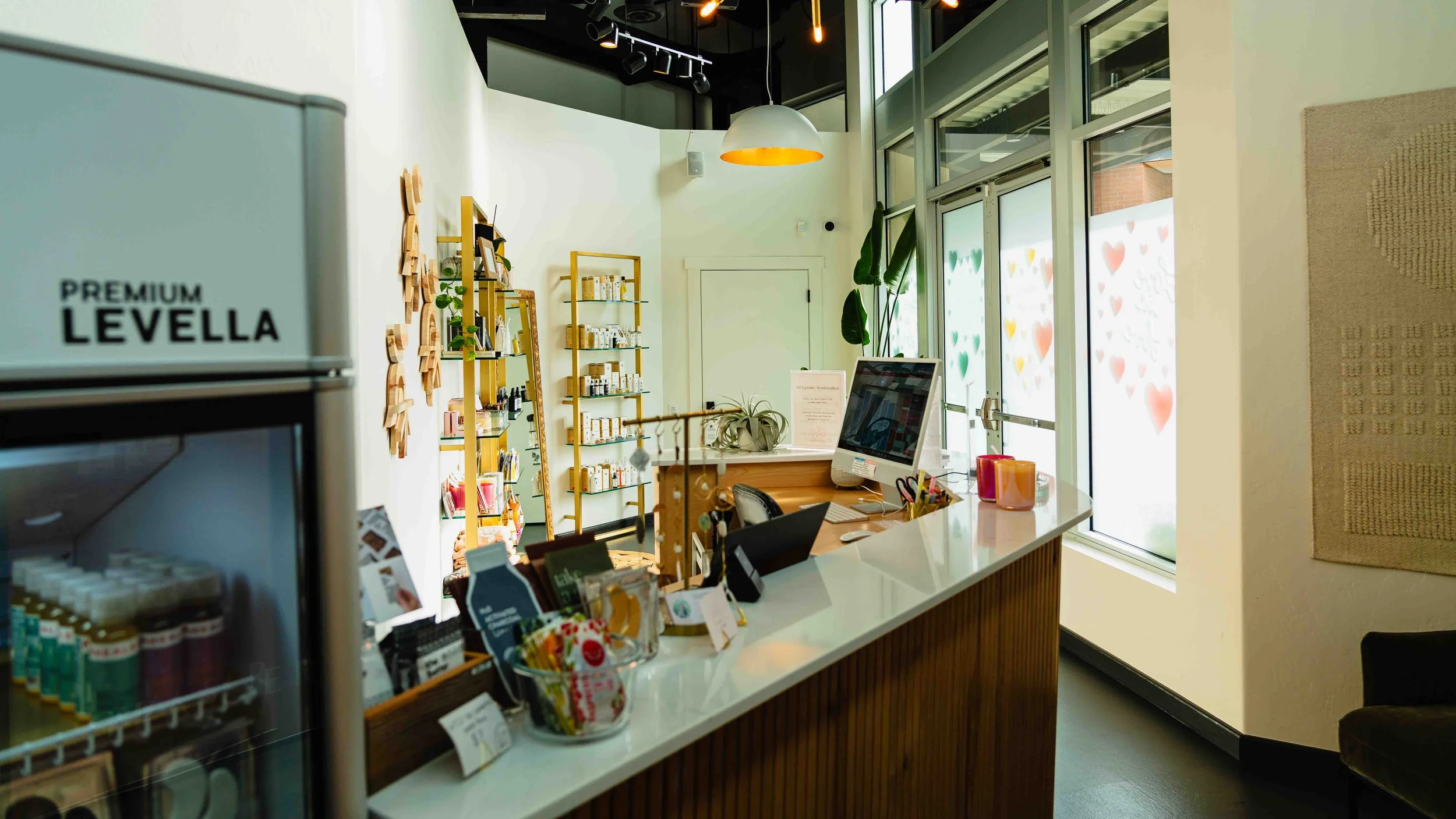 View of a retail store or office with a counter, shelves with products, a computer on the counter, and large windows with decorative heart patterns, bright lighting, and a minimalistic interior design.
