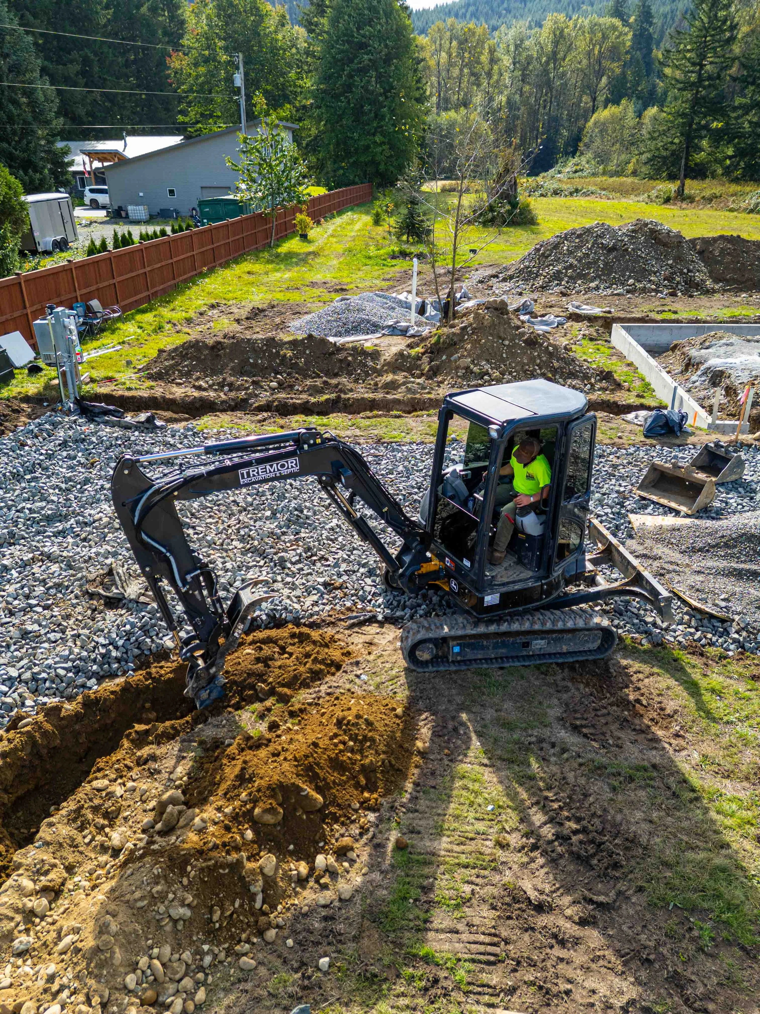 Excavator digging a trench in a yard with piles of dirt and gravel, surrounded by trees and a fence.