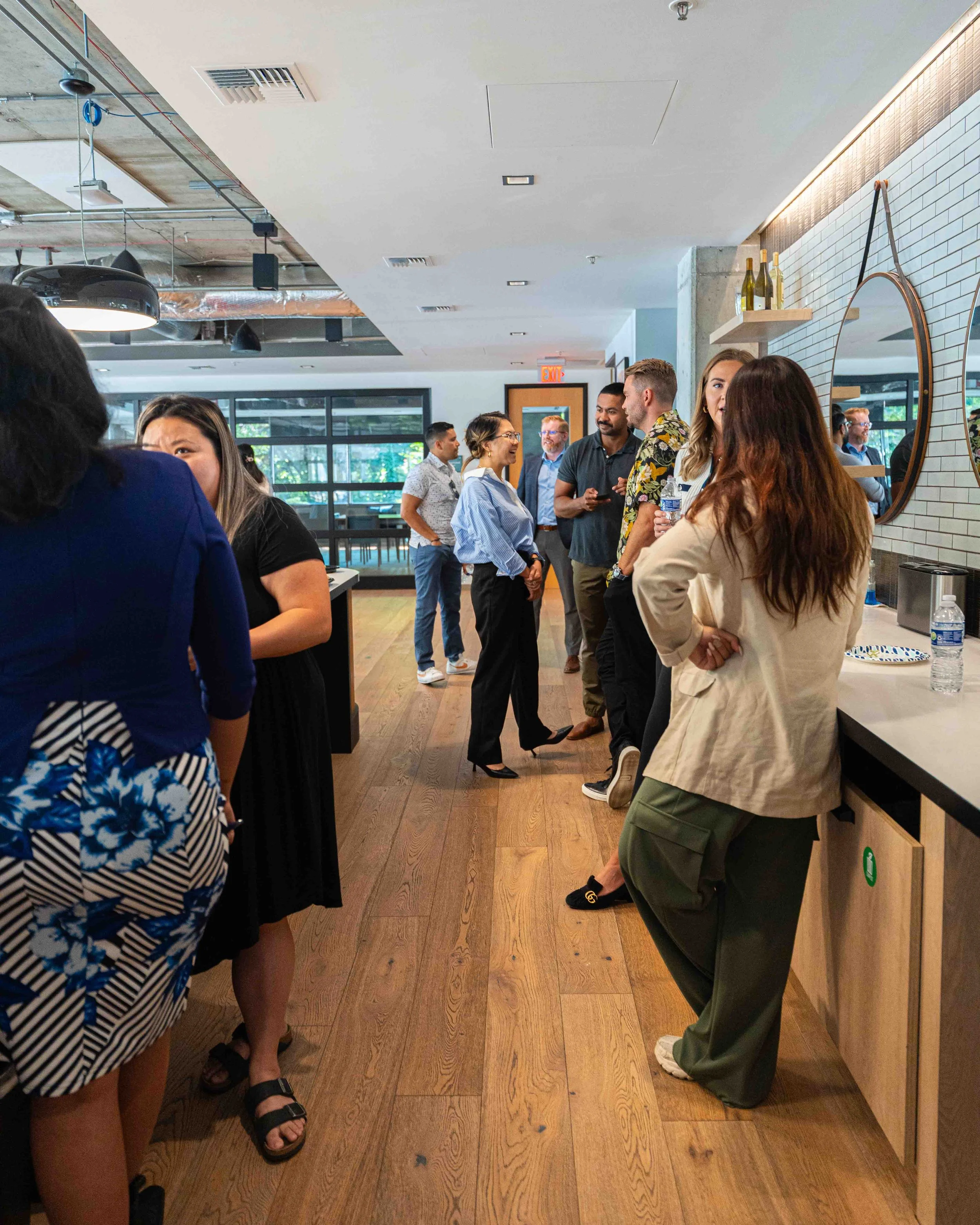 A group of diverse people engaged in conversation inside a modern, well-lit indoor space with wooden floors and white brick walls. Some are holding water bottles, and there are wine bottles on shelves and a mirror on the wall.