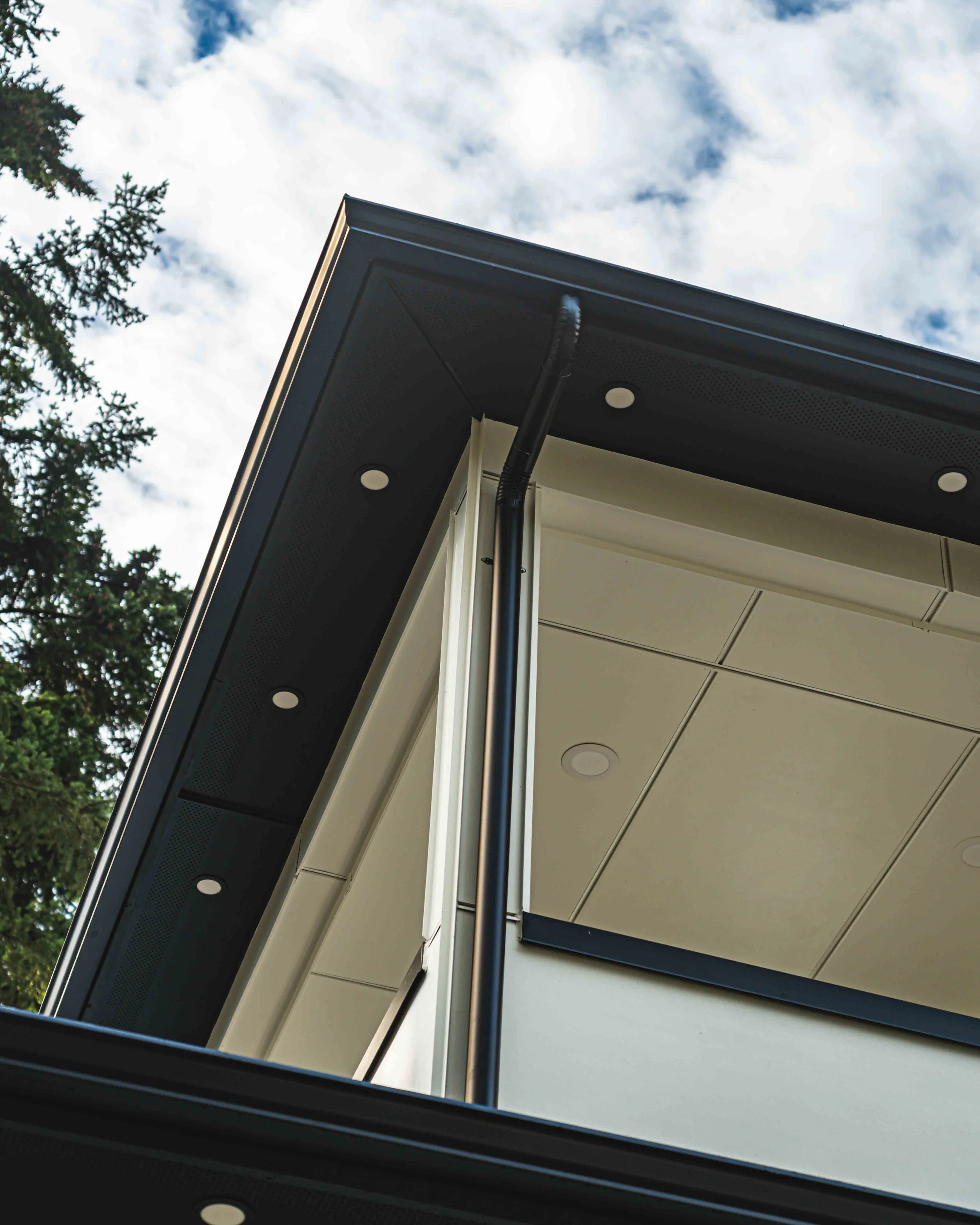Close-up of a modern building corner with black and white panels, a gutter pipe, and recessed lighting, against a partly cloudy sky with trees.