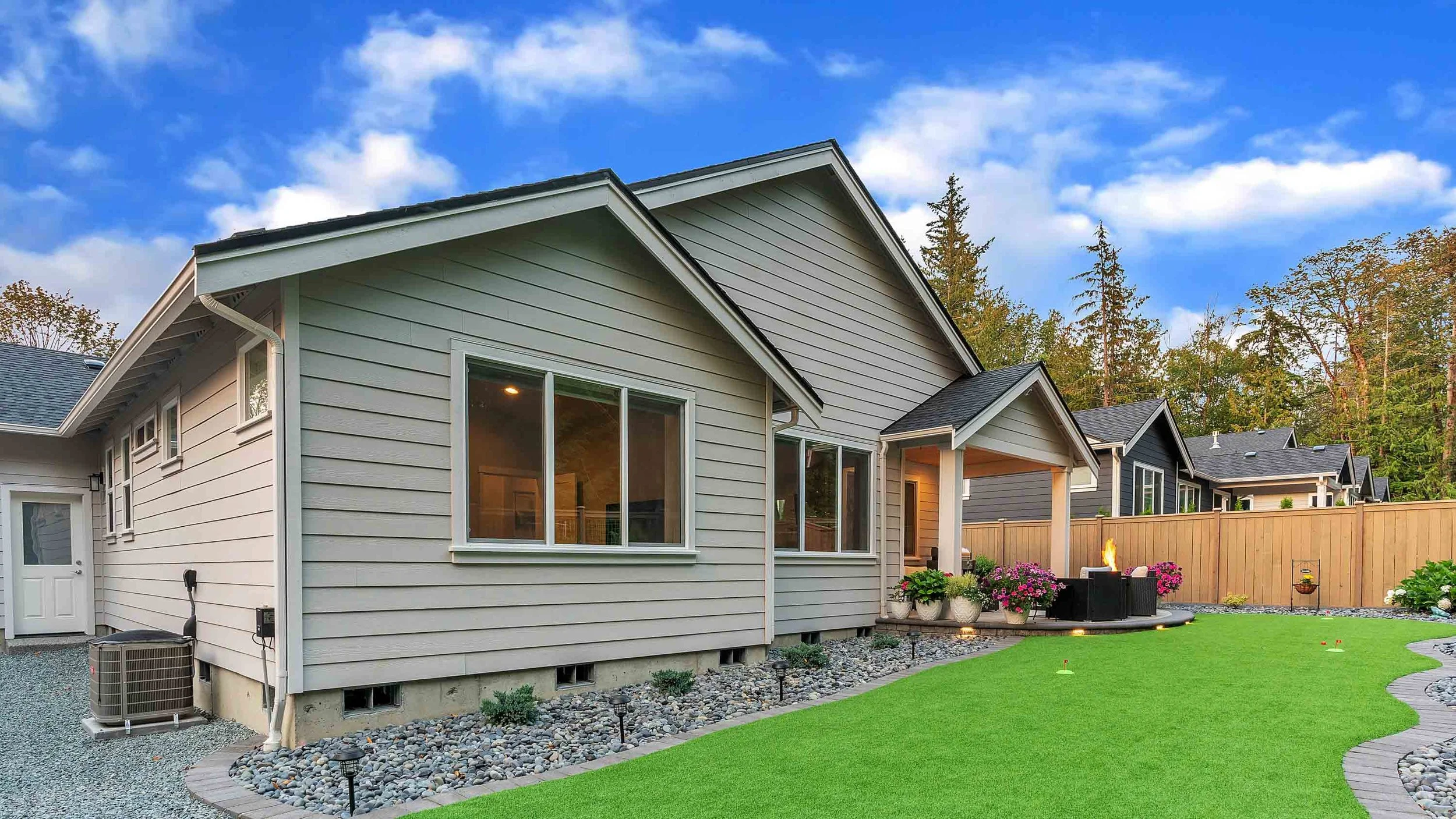 Backyard view of modern houses with beige and dark gray siding, large windows, a wooden fence, a small decorative garden, flowering plants, and a fire pit