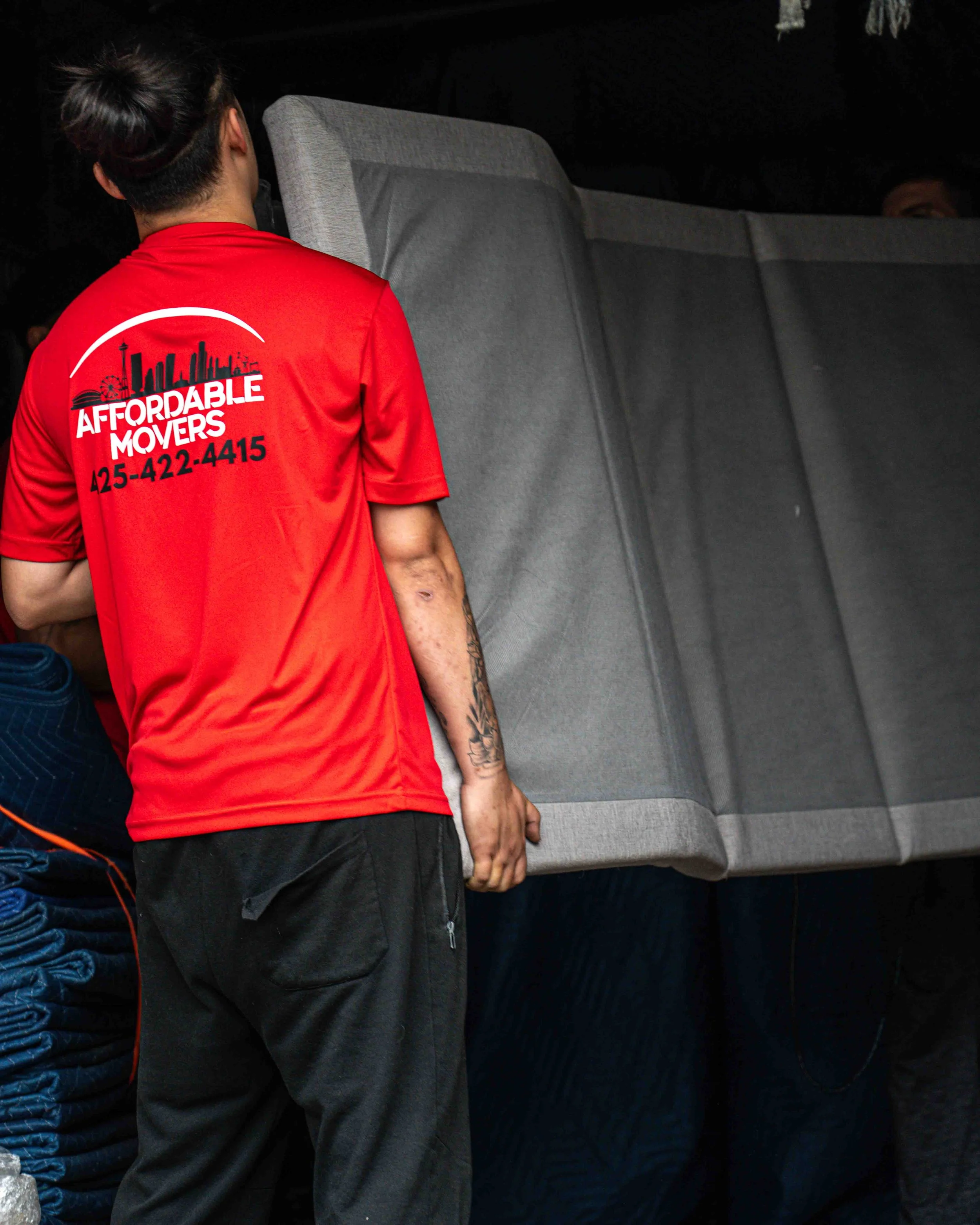 Person with tattooed arm in red t-shirt with 'Affordable Movers' logo and phone number, moving a piece of furniture in a dark room.