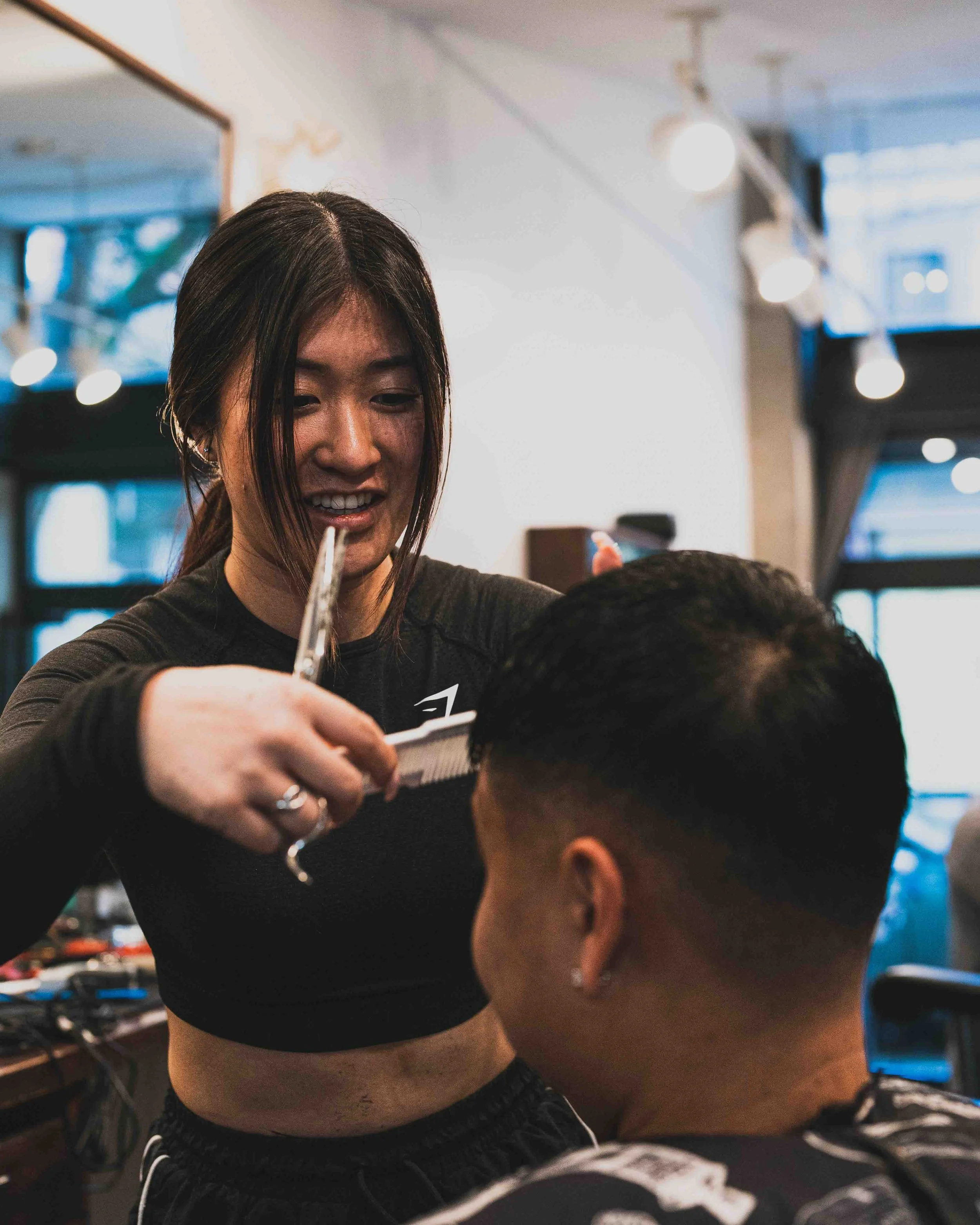 A hairstylist is cutting a person's hair in a salon. The stylist is smiling and appears focused on her task. The salon has modern lighting and decor, with blurred background elements including windows and salon equipment.