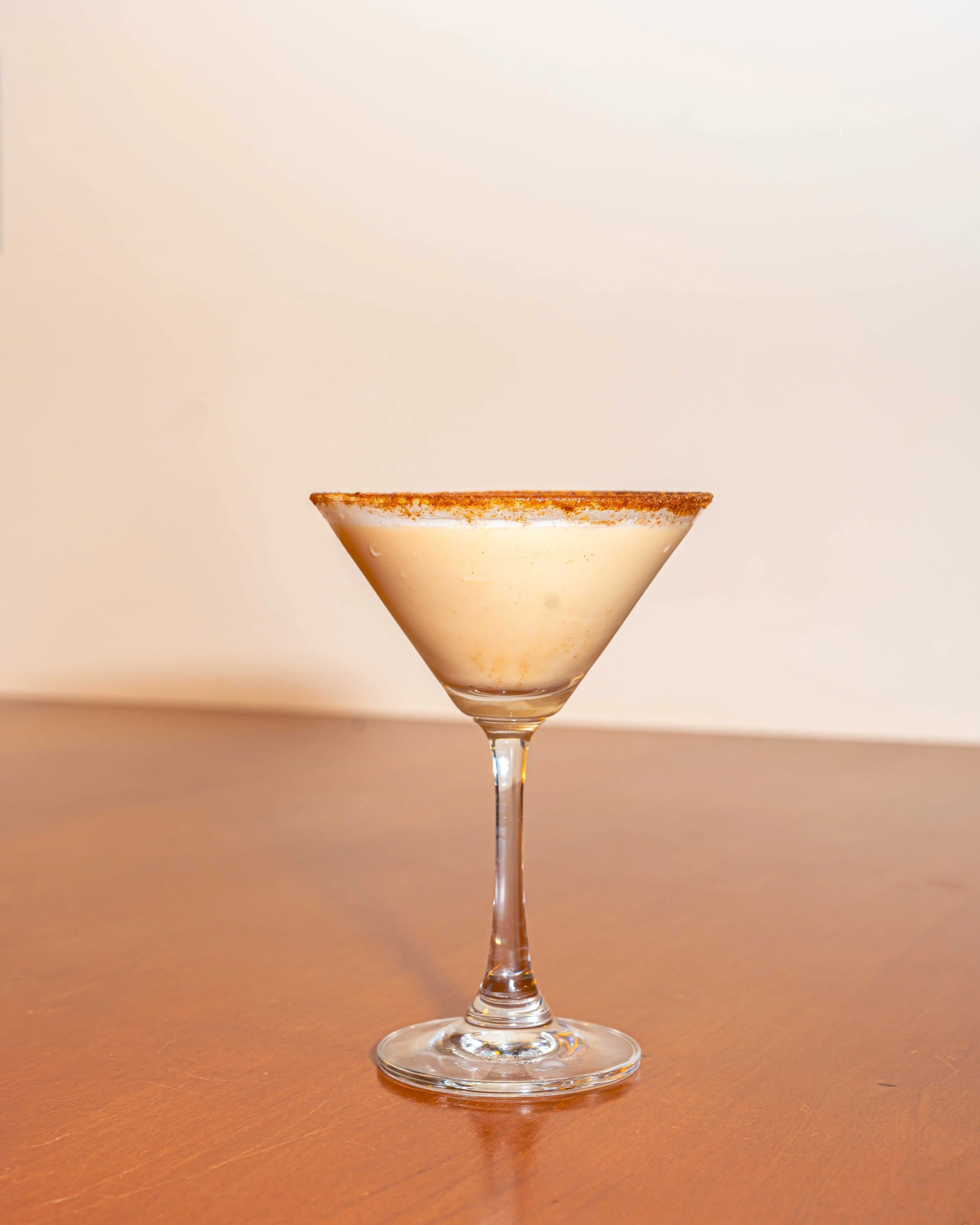 A cocktail glass with a frothy, cream-colored drink, garnished with a cinnamon or cocoa powder rim, on a wooden surface against a plain background.