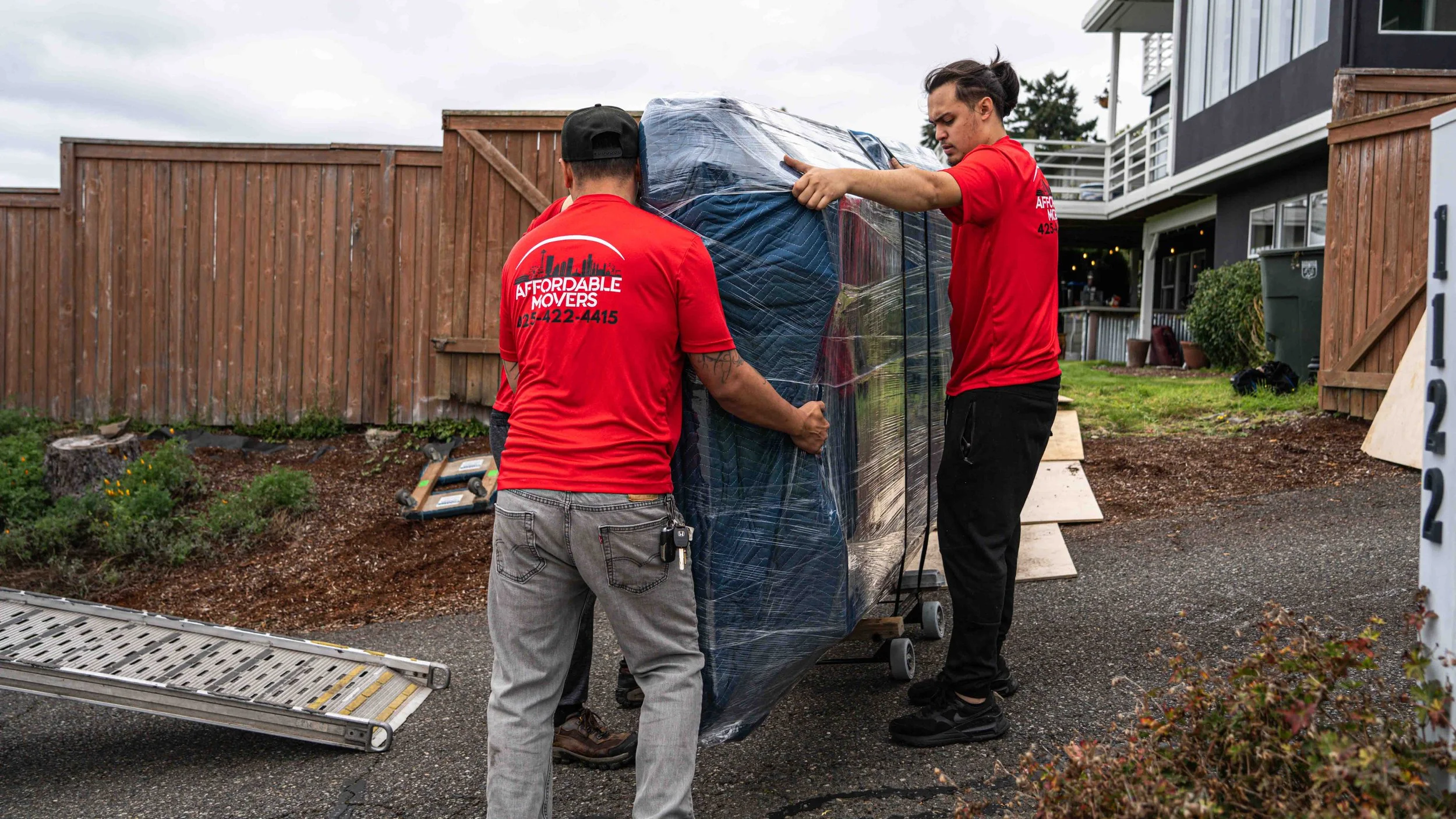 Two movers in red shirts loading a wrapped large piece of furniture or appliance onto a truck with a ramp outside a residential area.