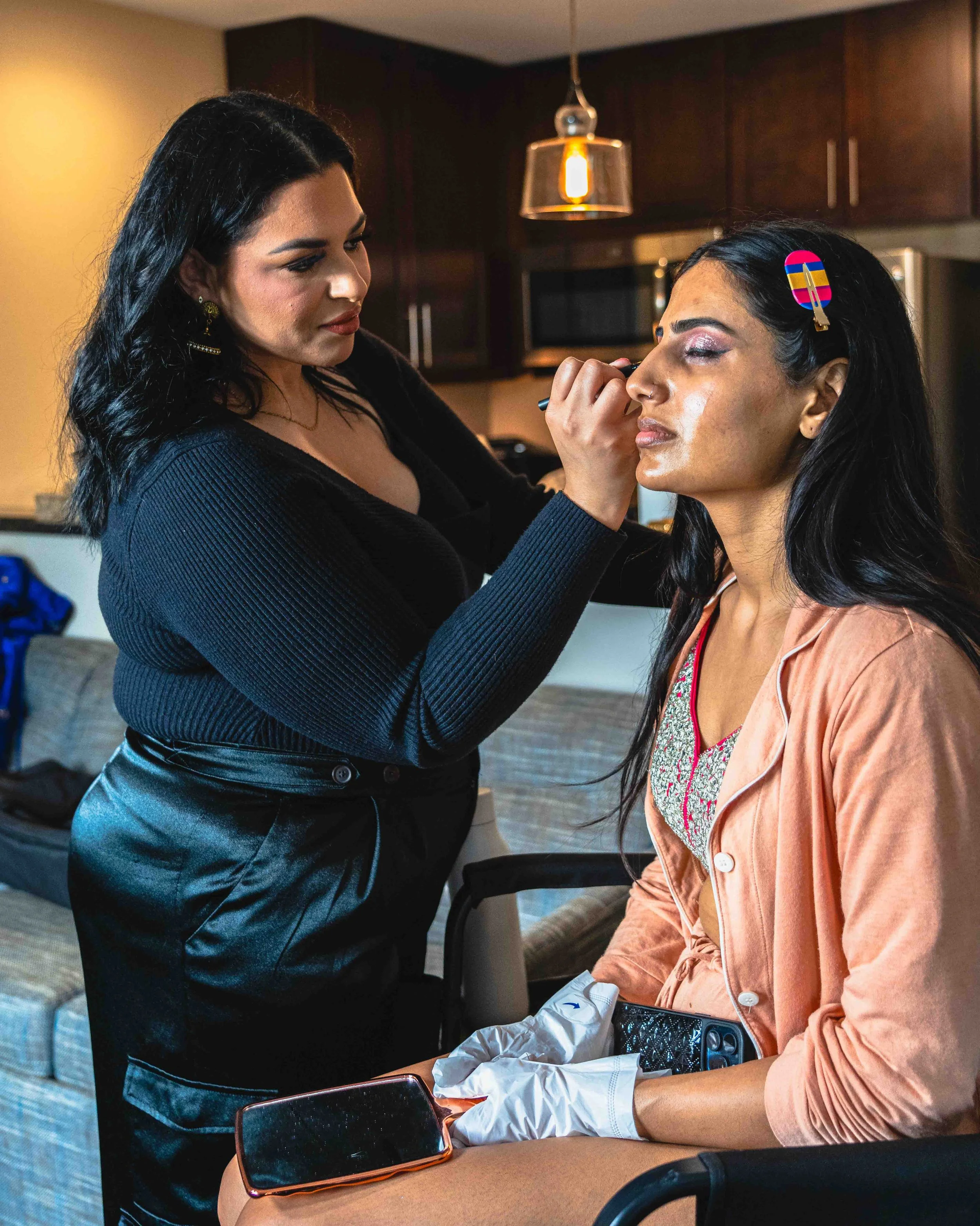 A woman with dark hair sitting at a table while another woman with black hair, face makeup, and colorful hair clip applies makeup around her eyes. The seated woman is wearing a peach-colored top and white gloves, with a makeup palette and smartphone 