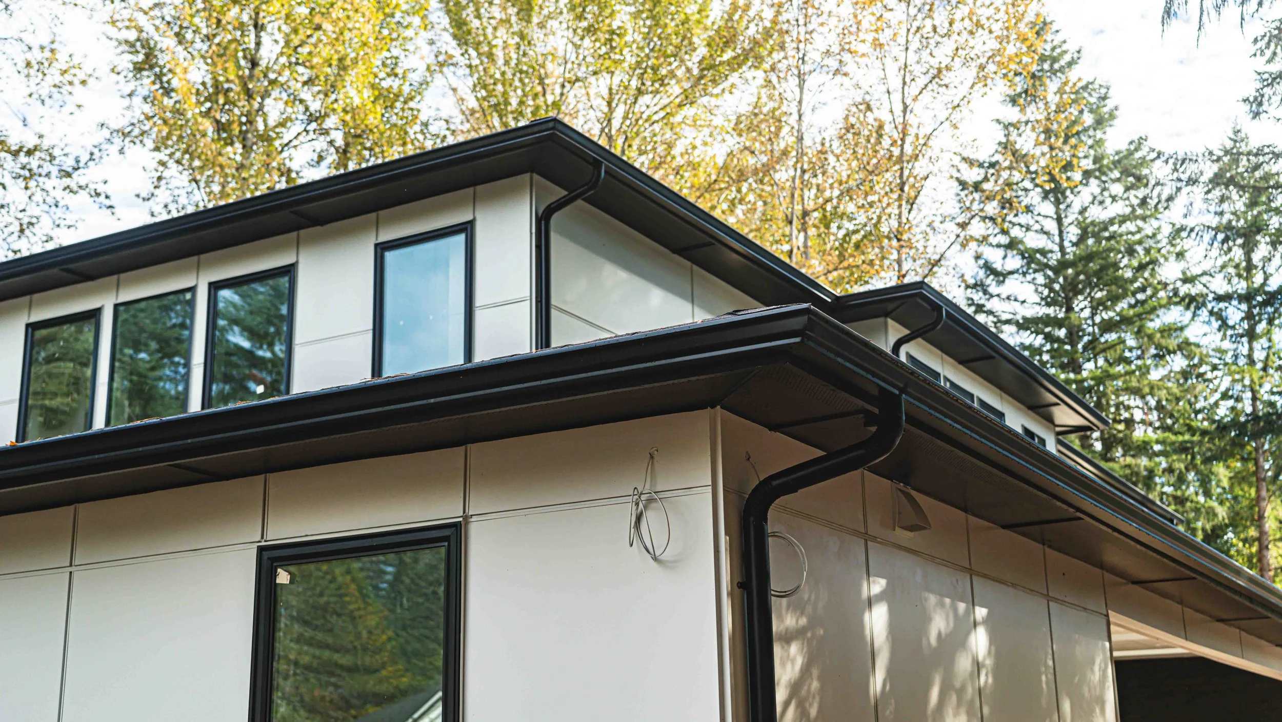 Close-up of the modern upper corner of a house with large windows, beige siding, black gutters, and trees with yellow and green leaves in the background.
