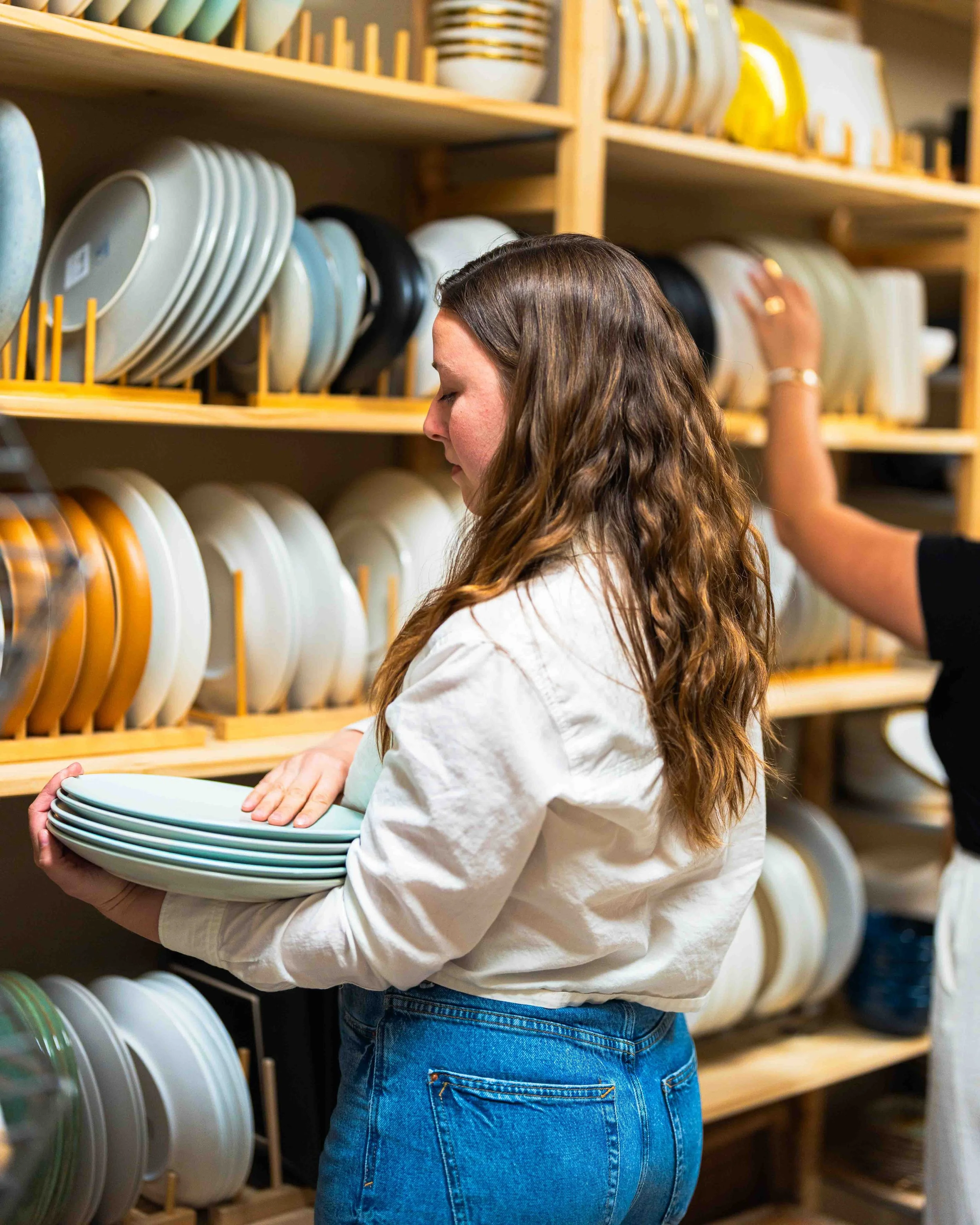 A woman with long, wavy brown hair wearing a white shirt and blue jeans is holding a stack of plates in a dishware store. She is facing shelves filled with various plates and dishes of different colors and patterns.