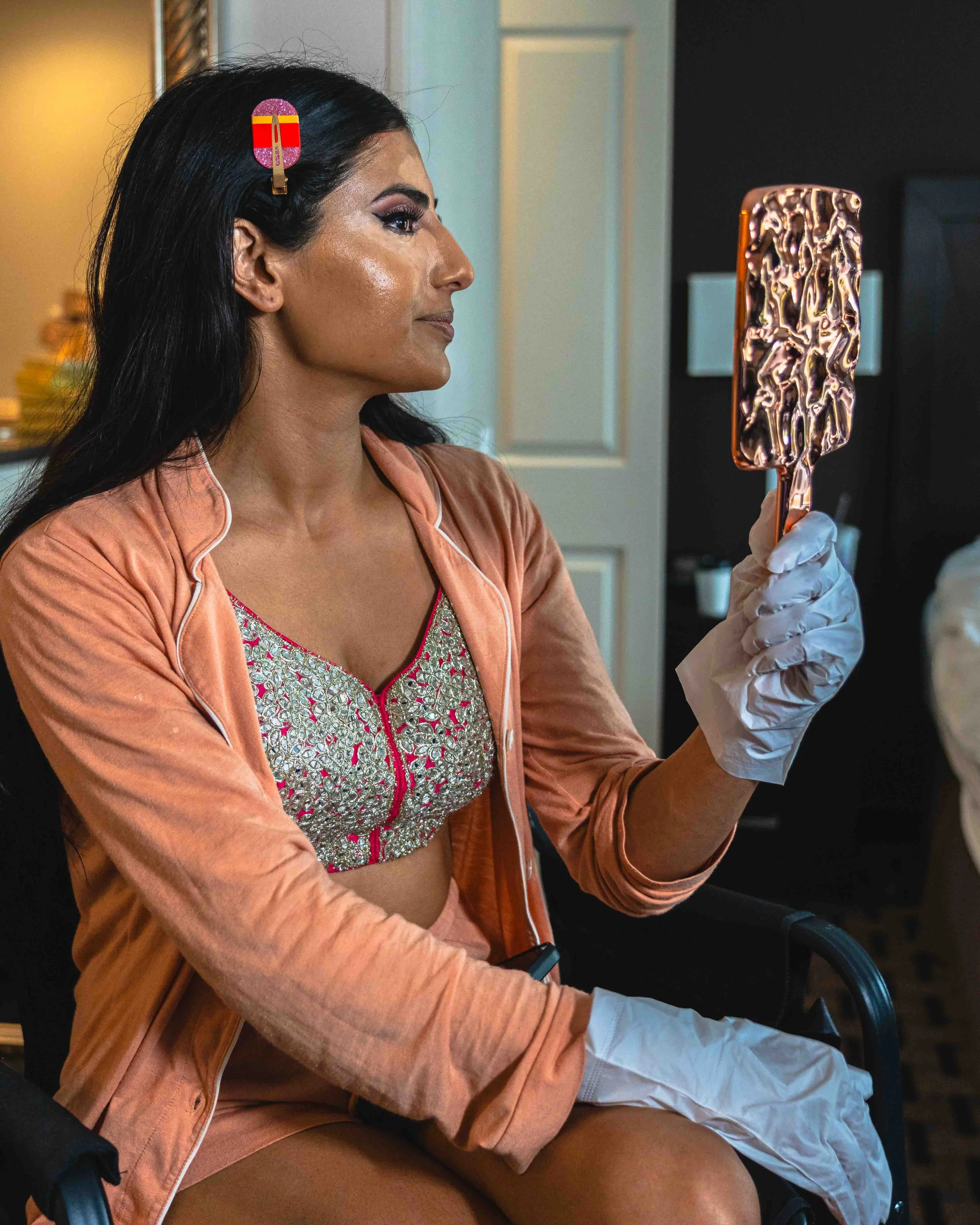 A woman with dark hair, wearing makeup, a peach-colored jacket, and a sparkly top, is sitting in a wheelchair. She is holding a shiny, textured copper-colored mirror in her right hand and looking at her reflection.