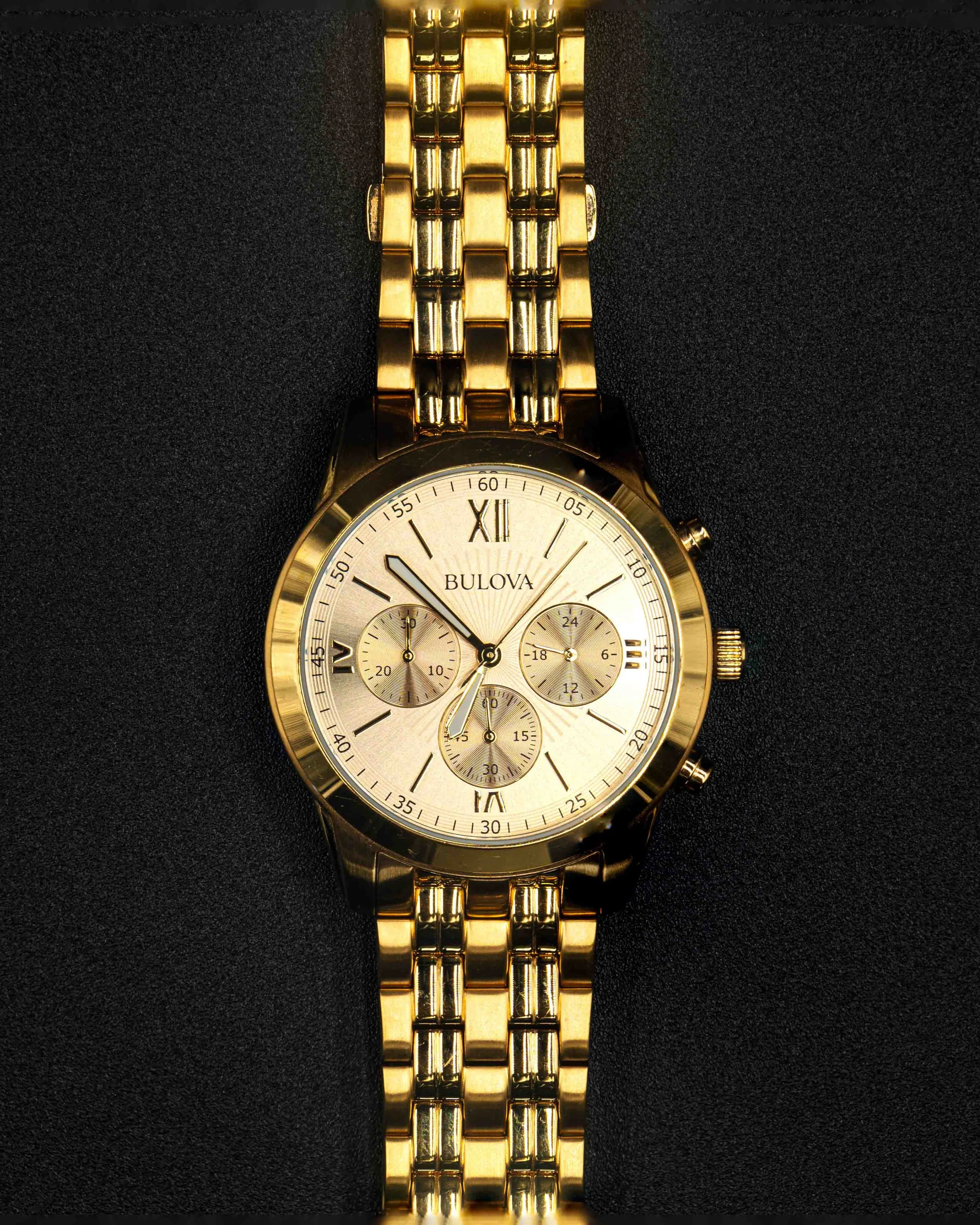 Close-up of a gold Bulova wristwatch with a metal band, displaying the time on a beige dial with Roman numeral and index markers, set against a black background.