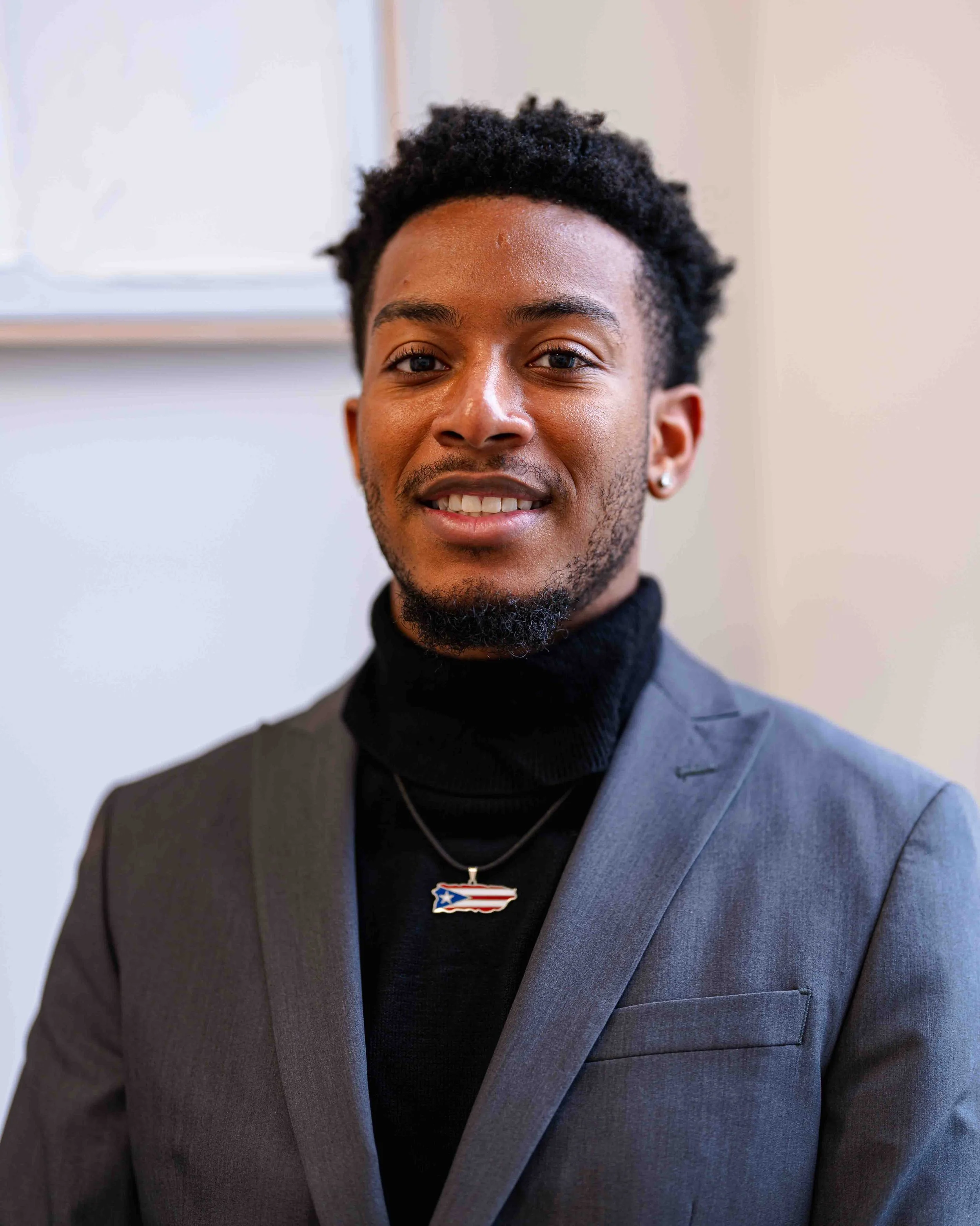 Close-up of a young man with dark curly hair, wearing a grey suit, black turtleneck, and a necklace with a pendant shaped like the state of North Carolina with the American flag inside. He is smiling slightly, and is standing in front of a plain, lig