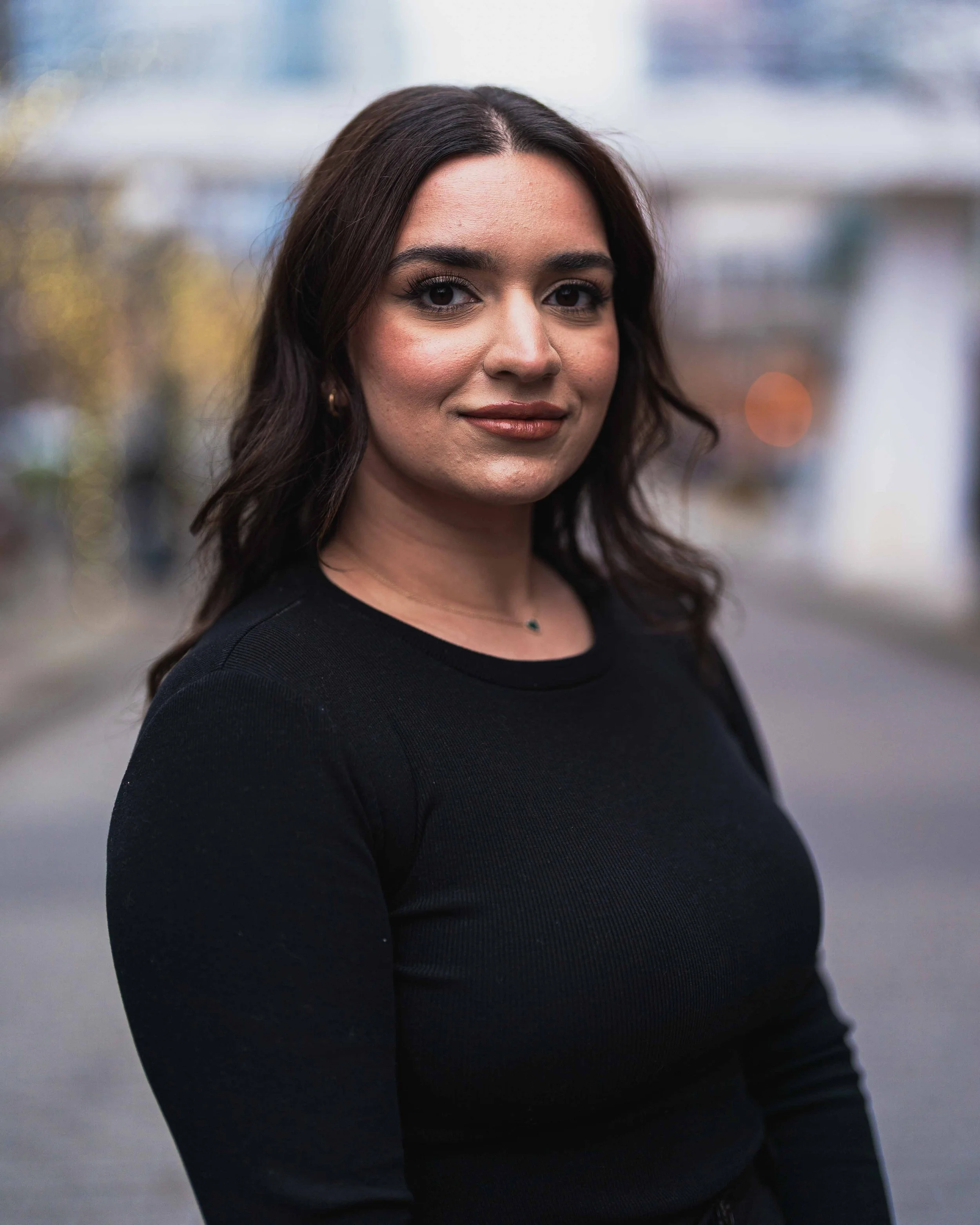 A woman with long dark hair, wearing makeup, a black top, and a necklace, standing outdoors with a blurred city background.
