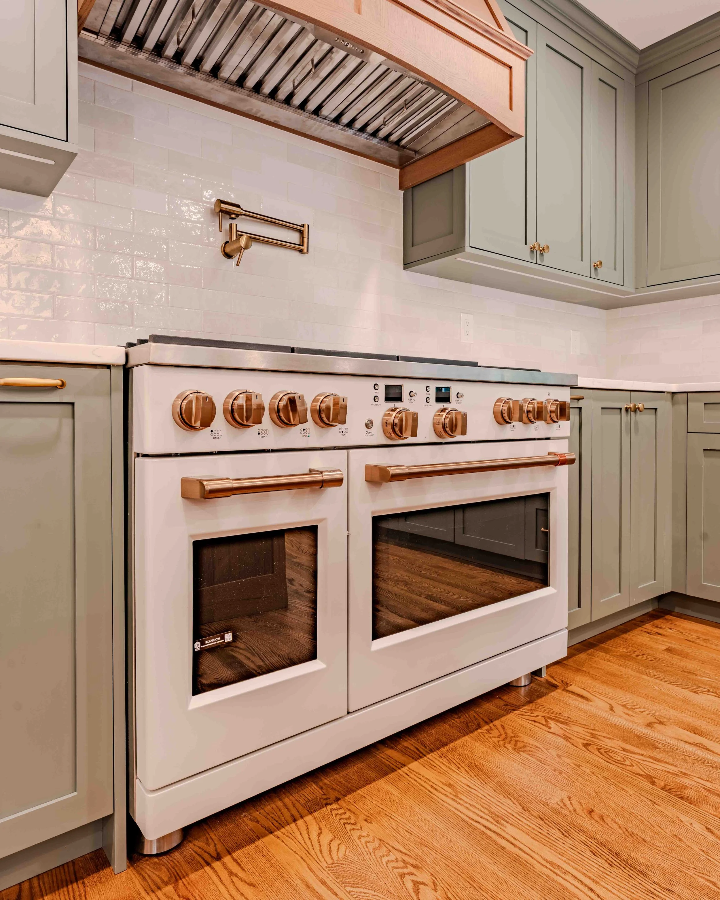 Modern kitchen with light green cabinets, a white oven with copper knobs, a brass pot filler above the stove, and wood flooring.