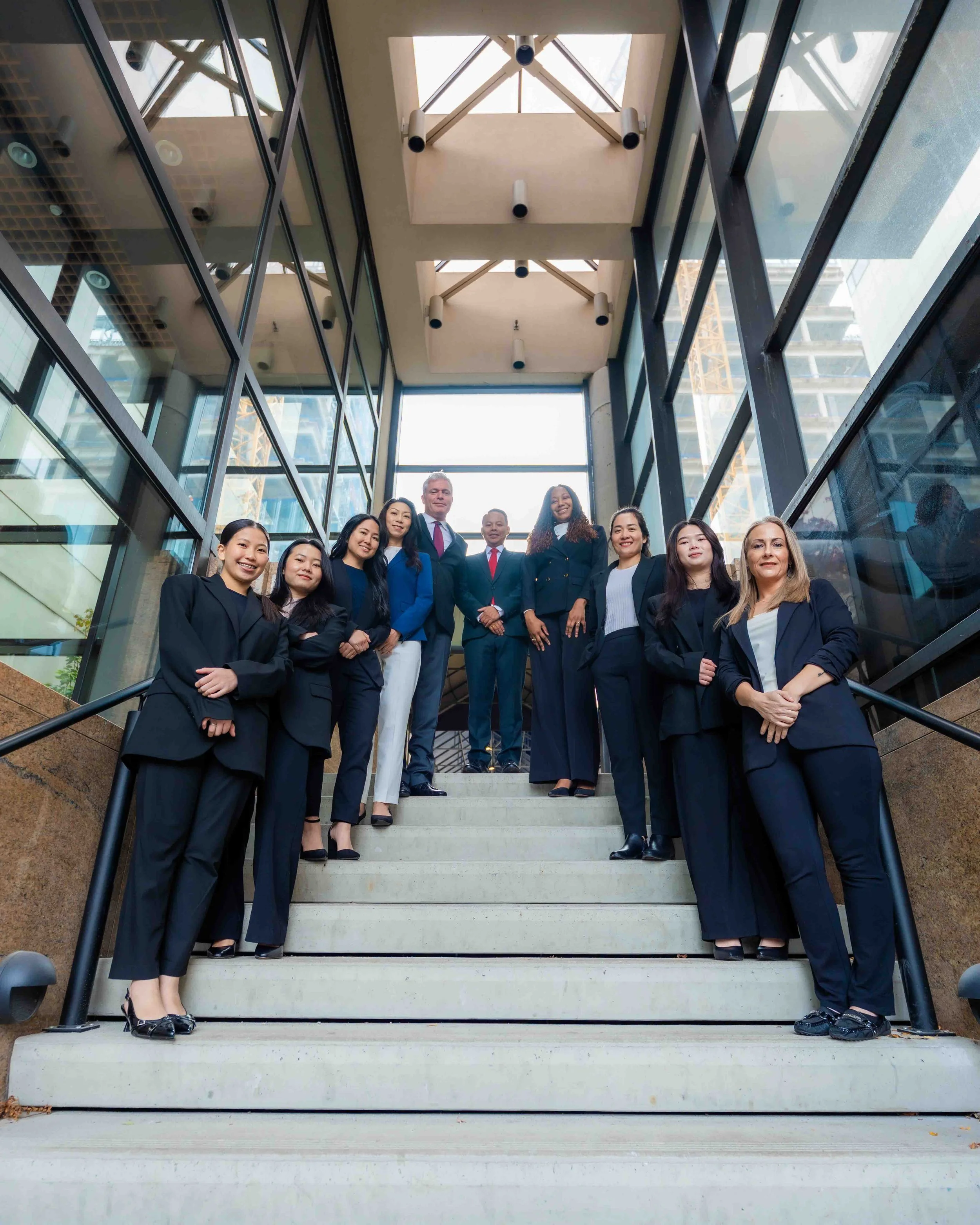 Group of ten people in business attire standing on a staircase inside a modern building with large glass windows and steel framing, smiling and looking at the camera.