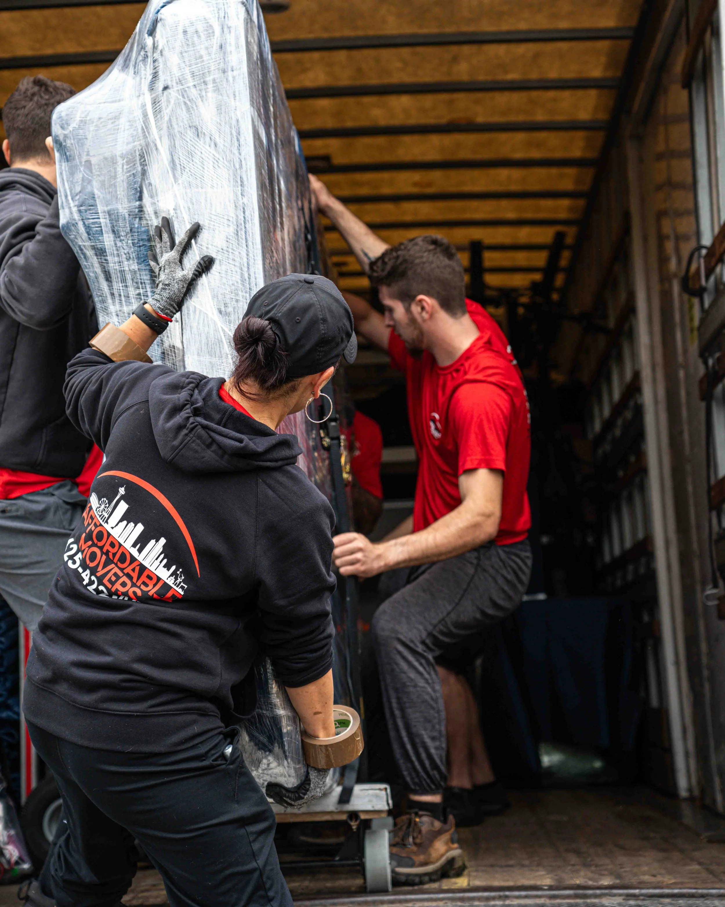 A group of people loading a large object wrapped in plastic onto a truck. One person is holding a roll of tape, and others are helping lift or support the object.