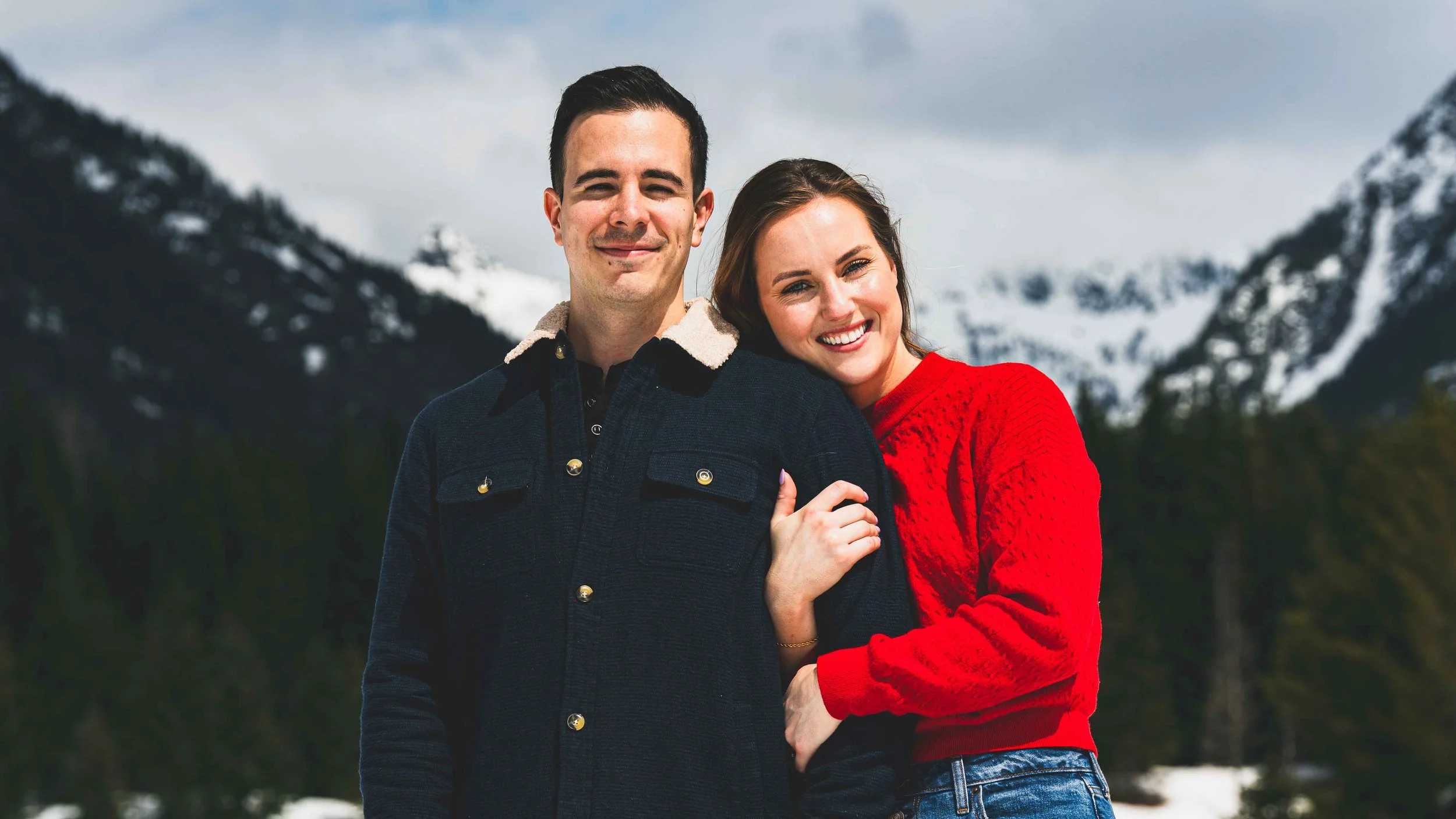 A smiling man and woman standing close together outdoors with snowy mountains in the background. The woman has her arm around the man's shoulder, both wearing casual sweaters.