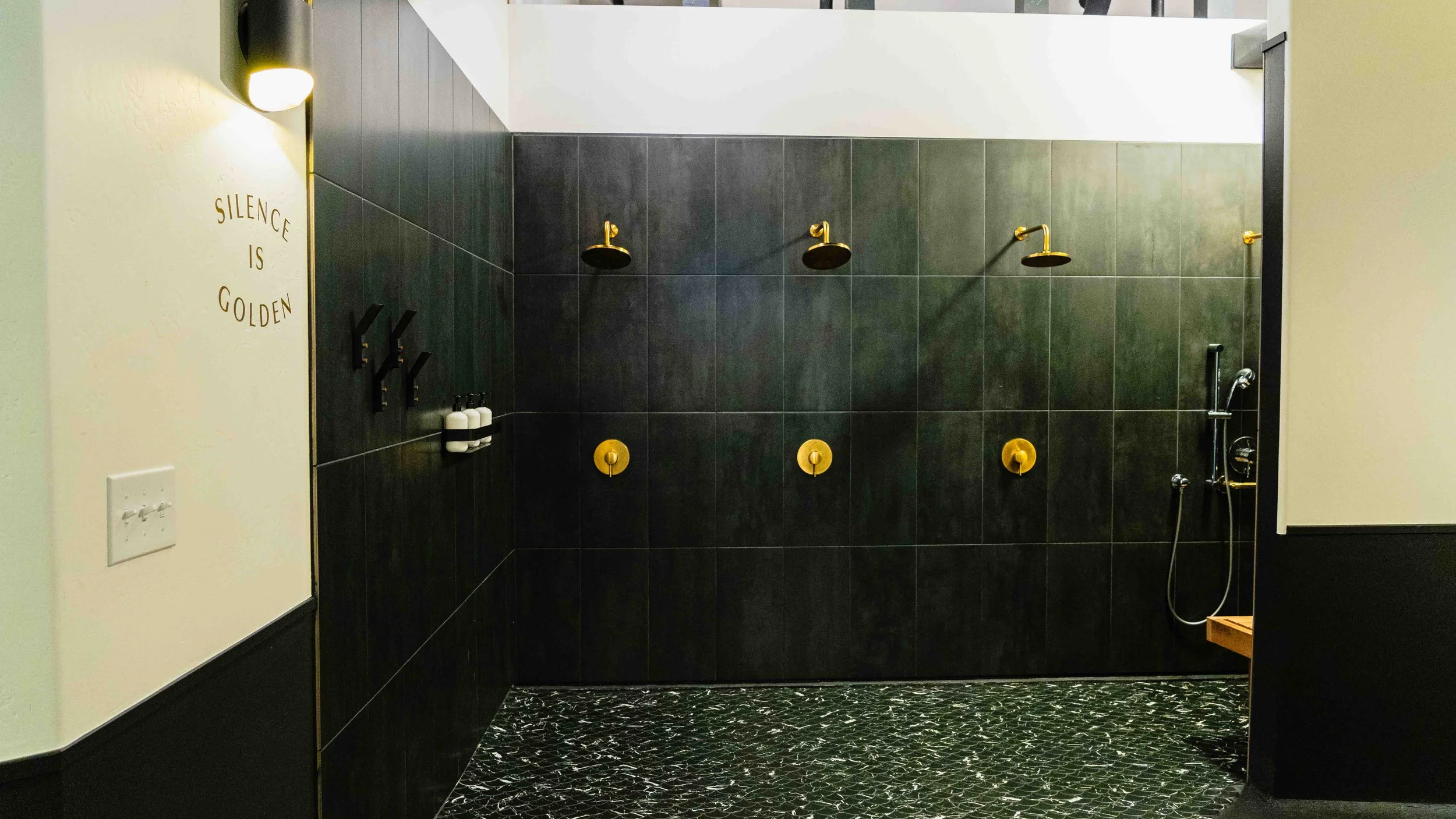 A modern shower area with black tiles, three gold shower heads, and black wall-mounted soap dispensers. The wall on the left has a sign reading 'Silence is Golden' and a light fixture. The floor has a black and white patterned design.