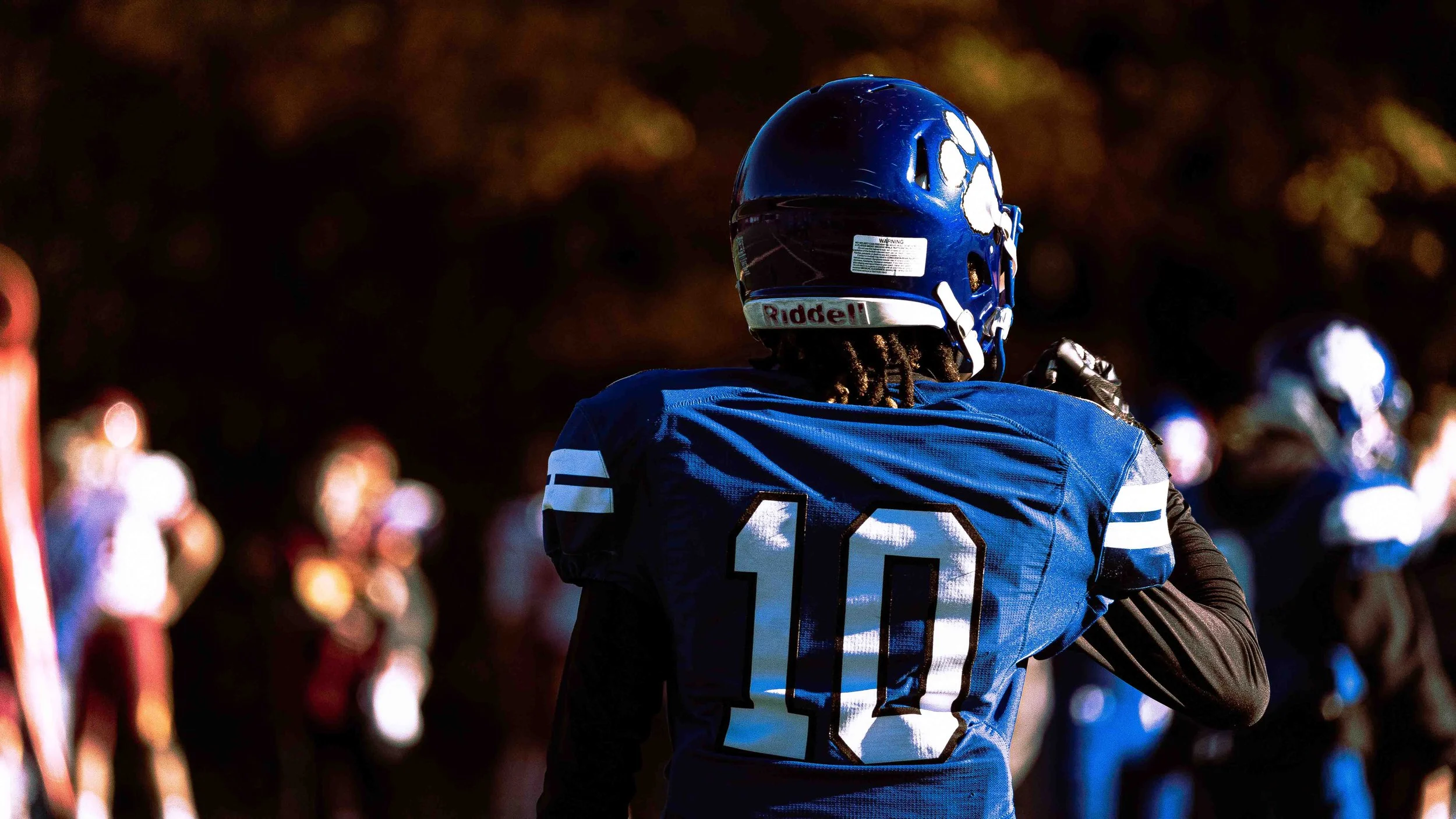 A football player wearing a blue helmet and jersey with the number 10 on the back, standing on the field during a game with other players and spectators in the background.