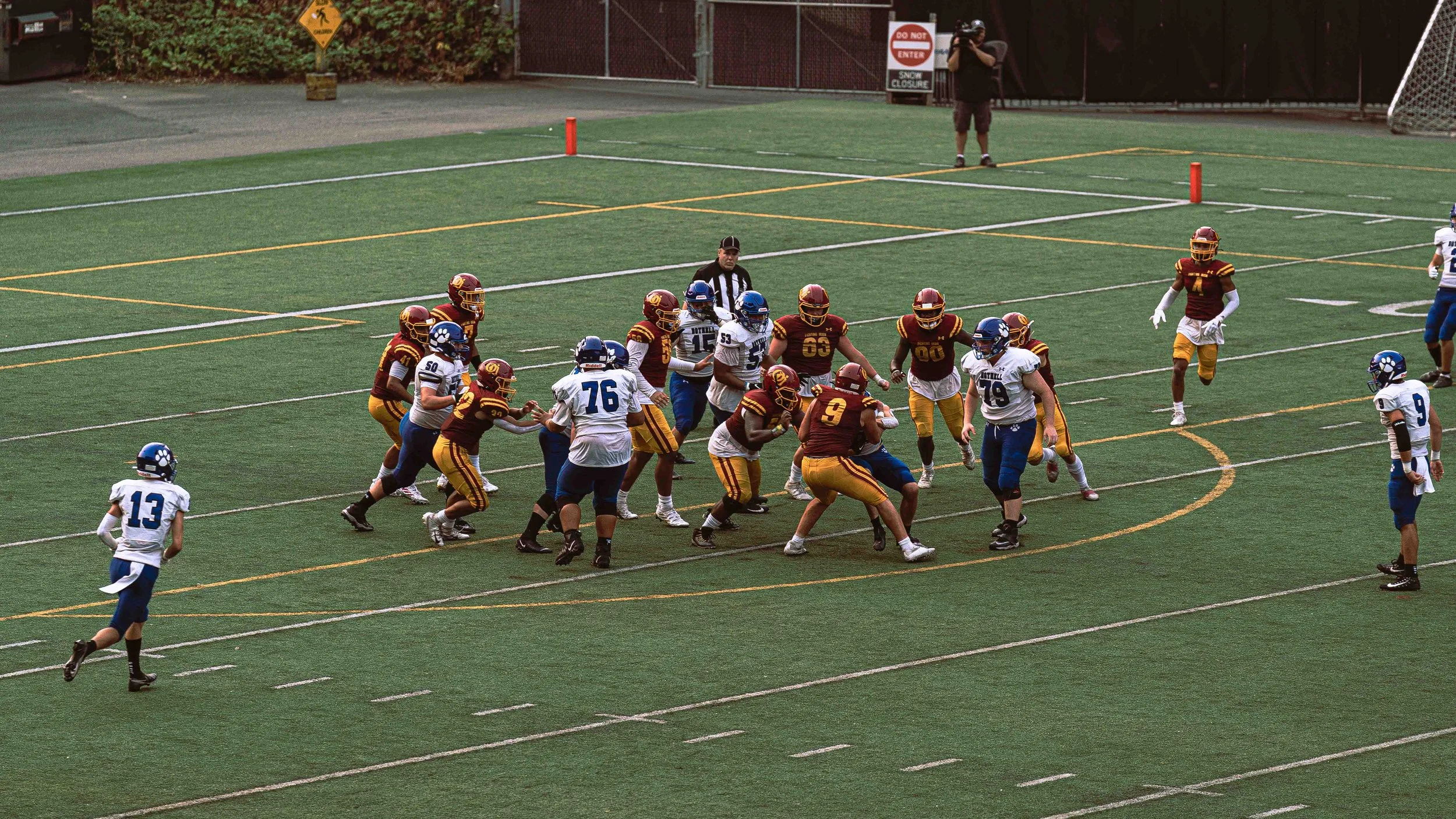 An American football game with players on the field, some in maroon and yellow uniforms and others in white and blue uniforms, with referees and spectators in the background.
