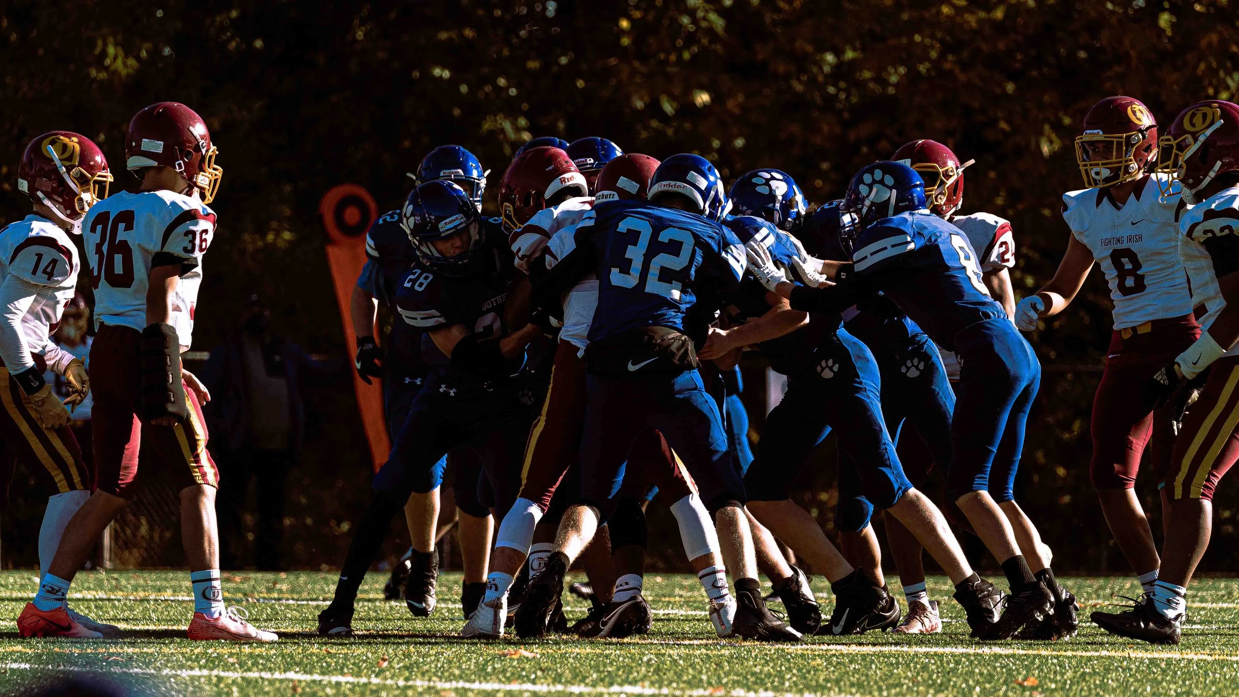 A close-up of a group of football players engaged in a play on the field, with some players wearing blue uniforms and others in white and red uniforms, during daytime with a blurry background of trees.