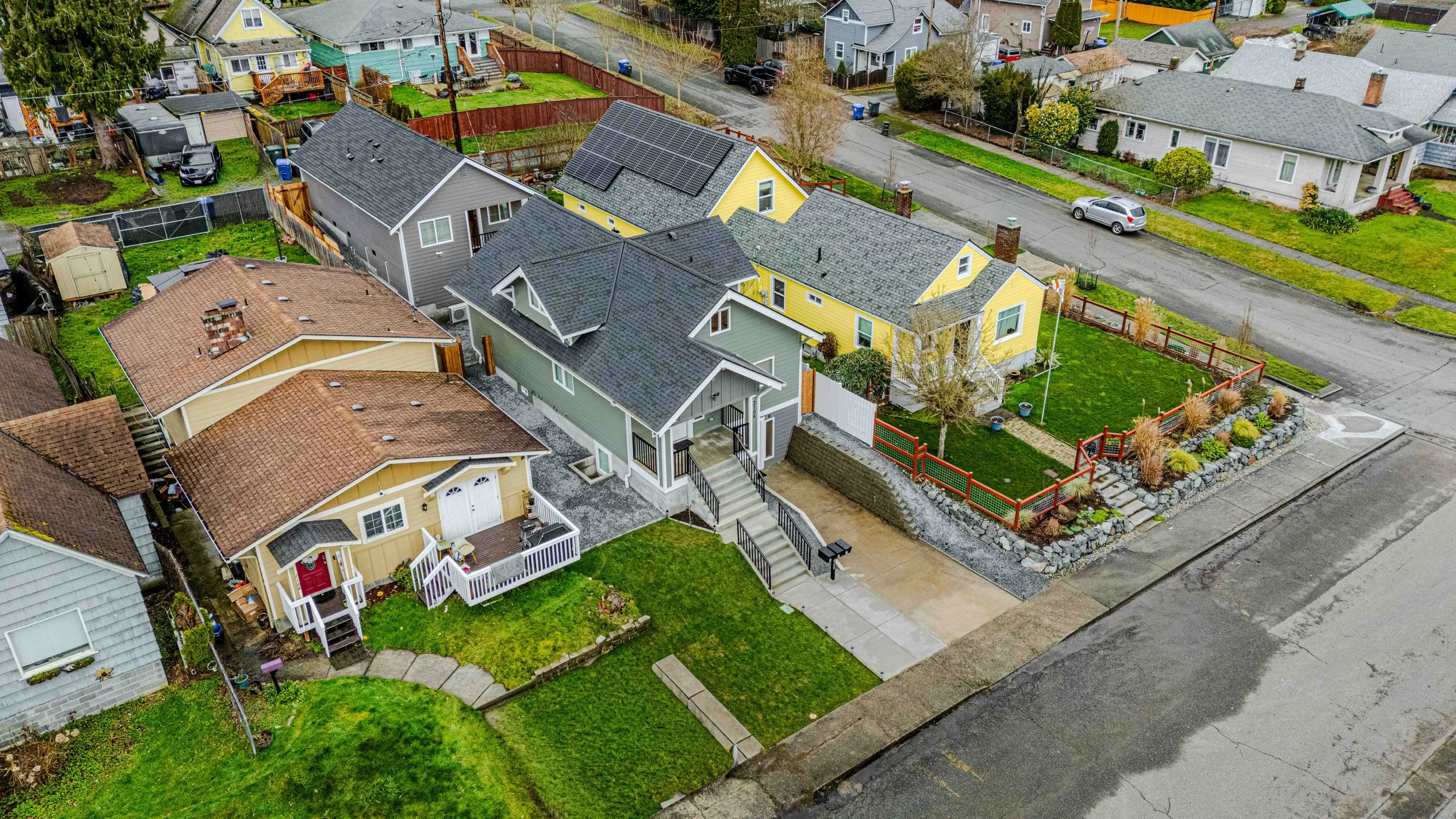 Aerial view of a residential neighborhood with multiple houses featuring different colored exteriors and yards, some with fences and gardens, and a street running through the neighborhood.